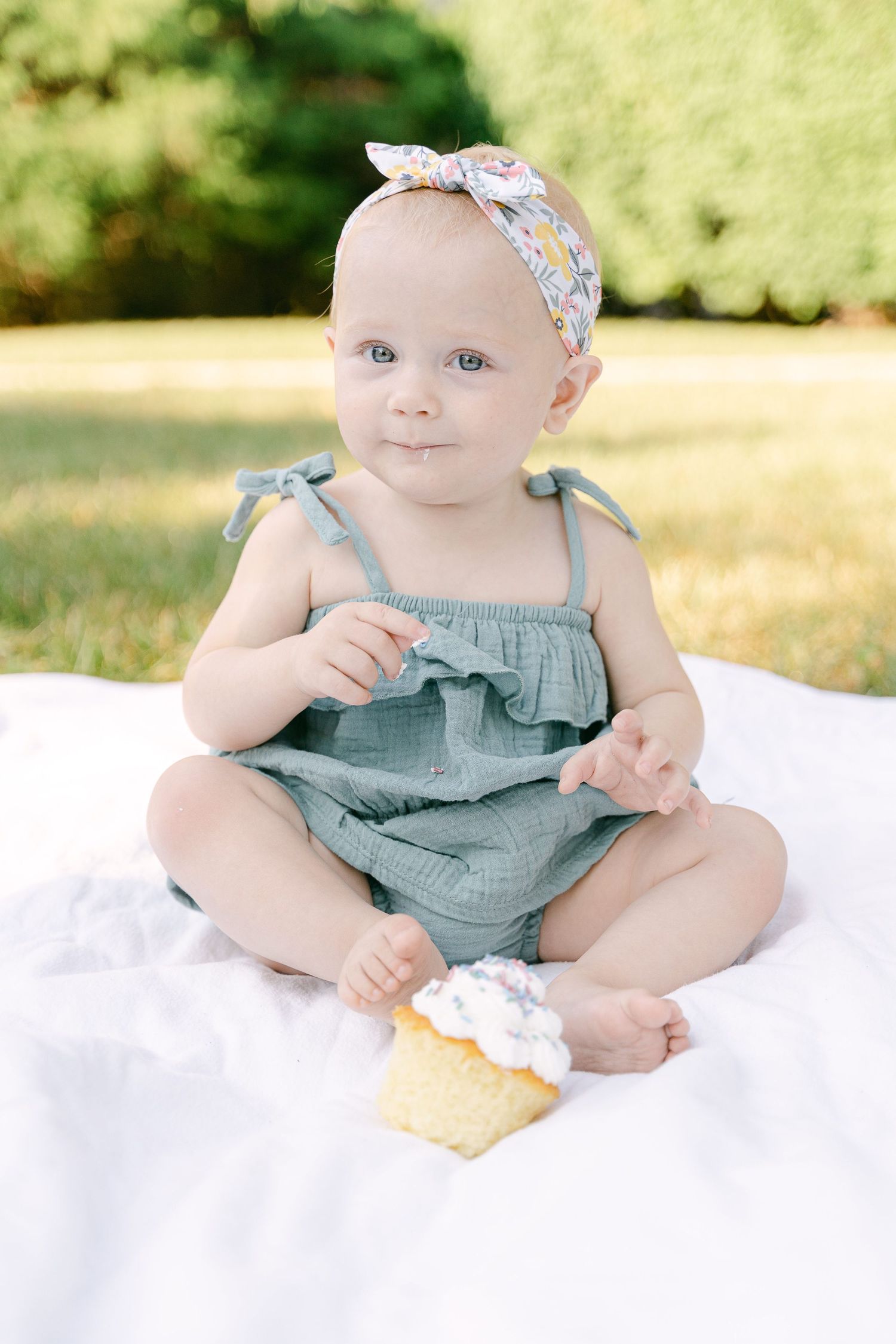 Baby in teal ruffled romper sits on white blanket with floral headband.