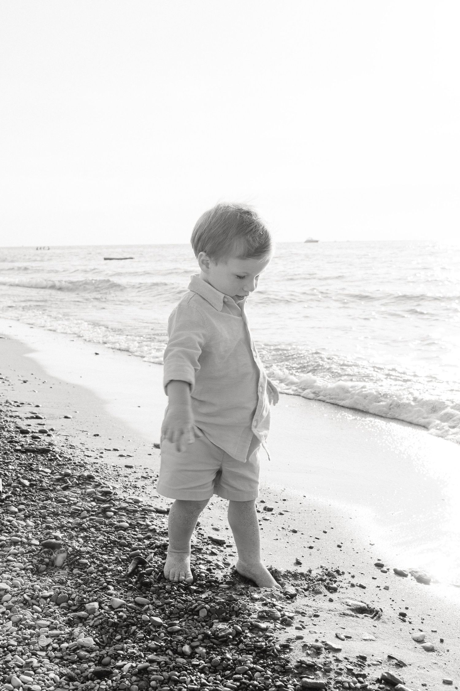 A toddler explores a rocky beach shoreline in black and white as gentle waves roll in.