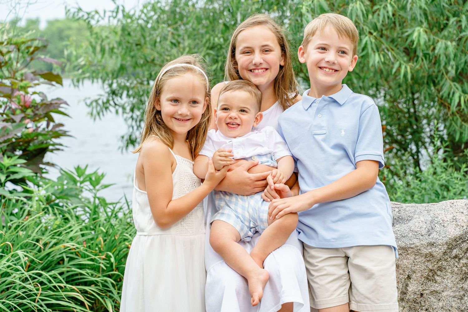 A group in coordinating white and blue outfits sits together on a log by the water during a summer photo session.