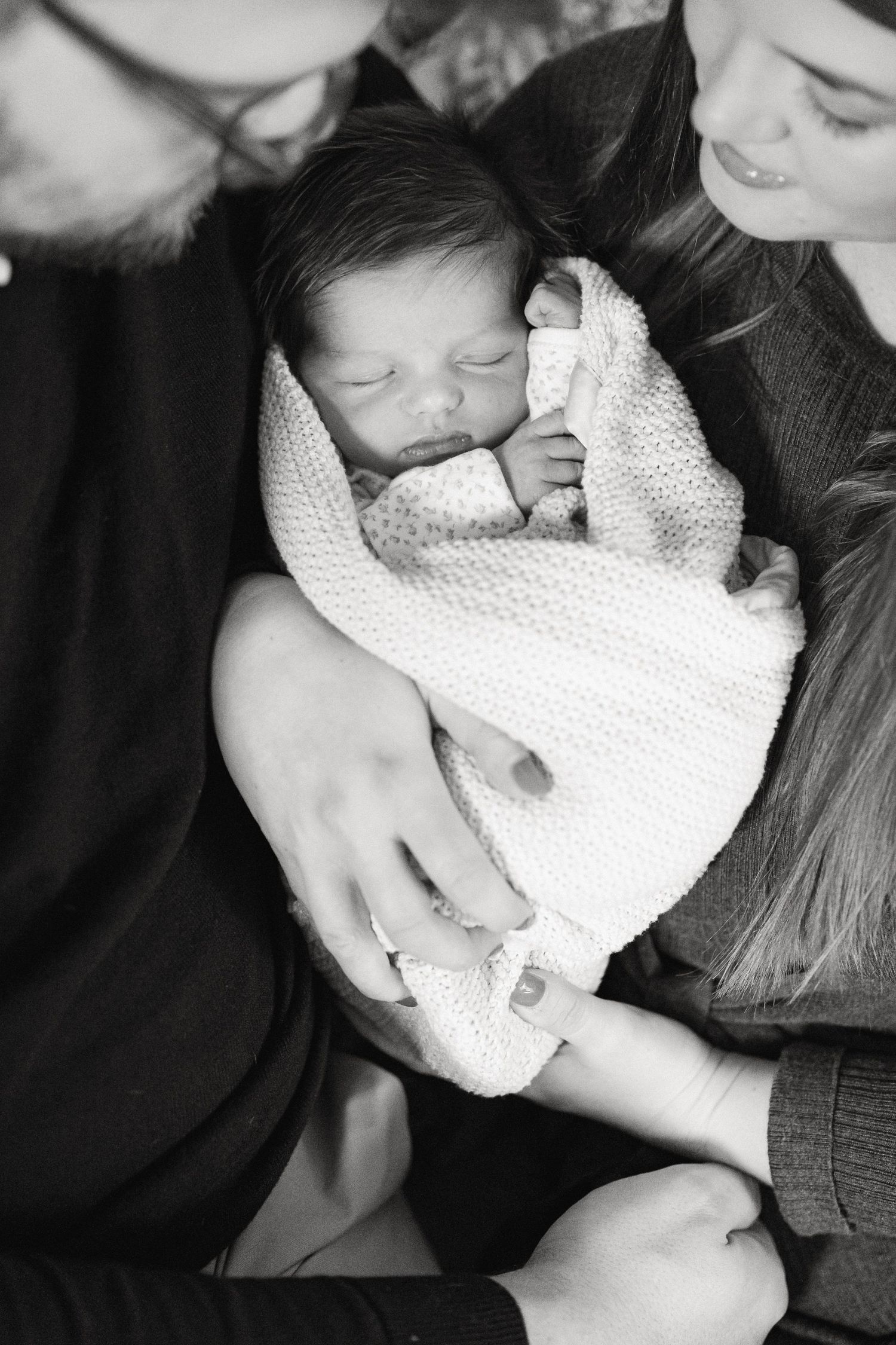 A tender black and white portrait of a newborn baby swaddled in a soft blanket being held by their parents.