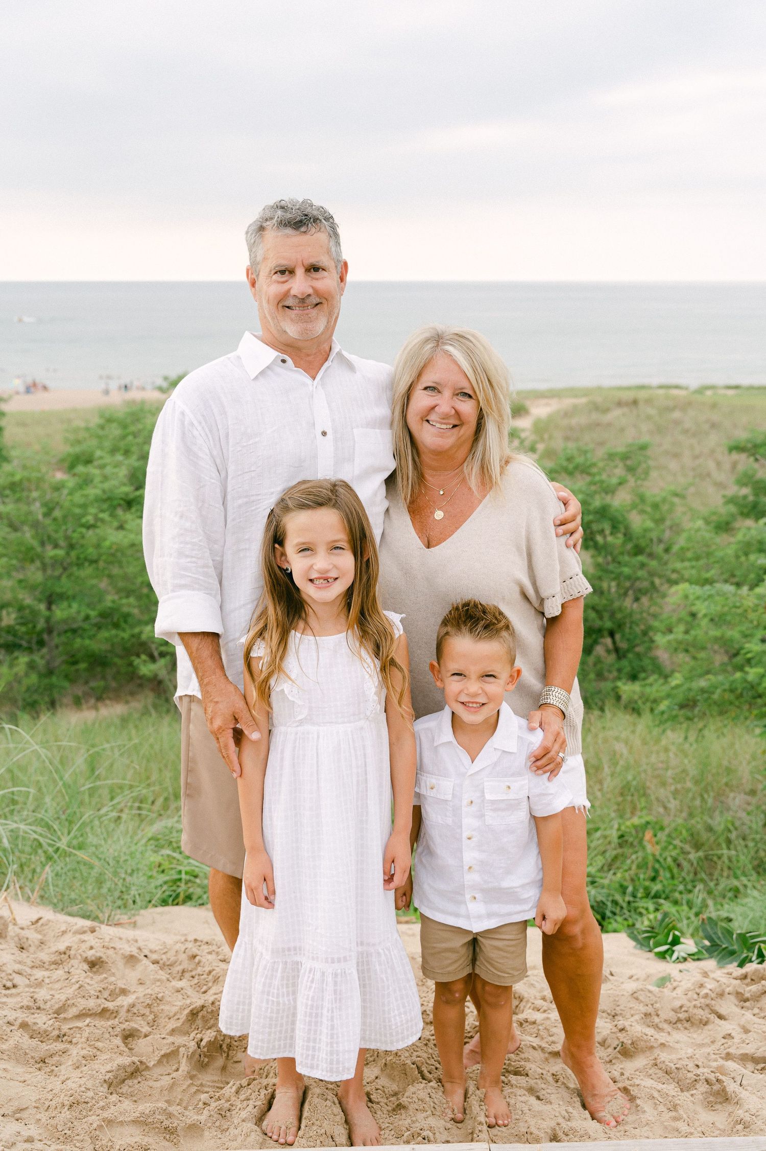 A family of four poses together on a sandy beach hill with the ocean visible in the background.