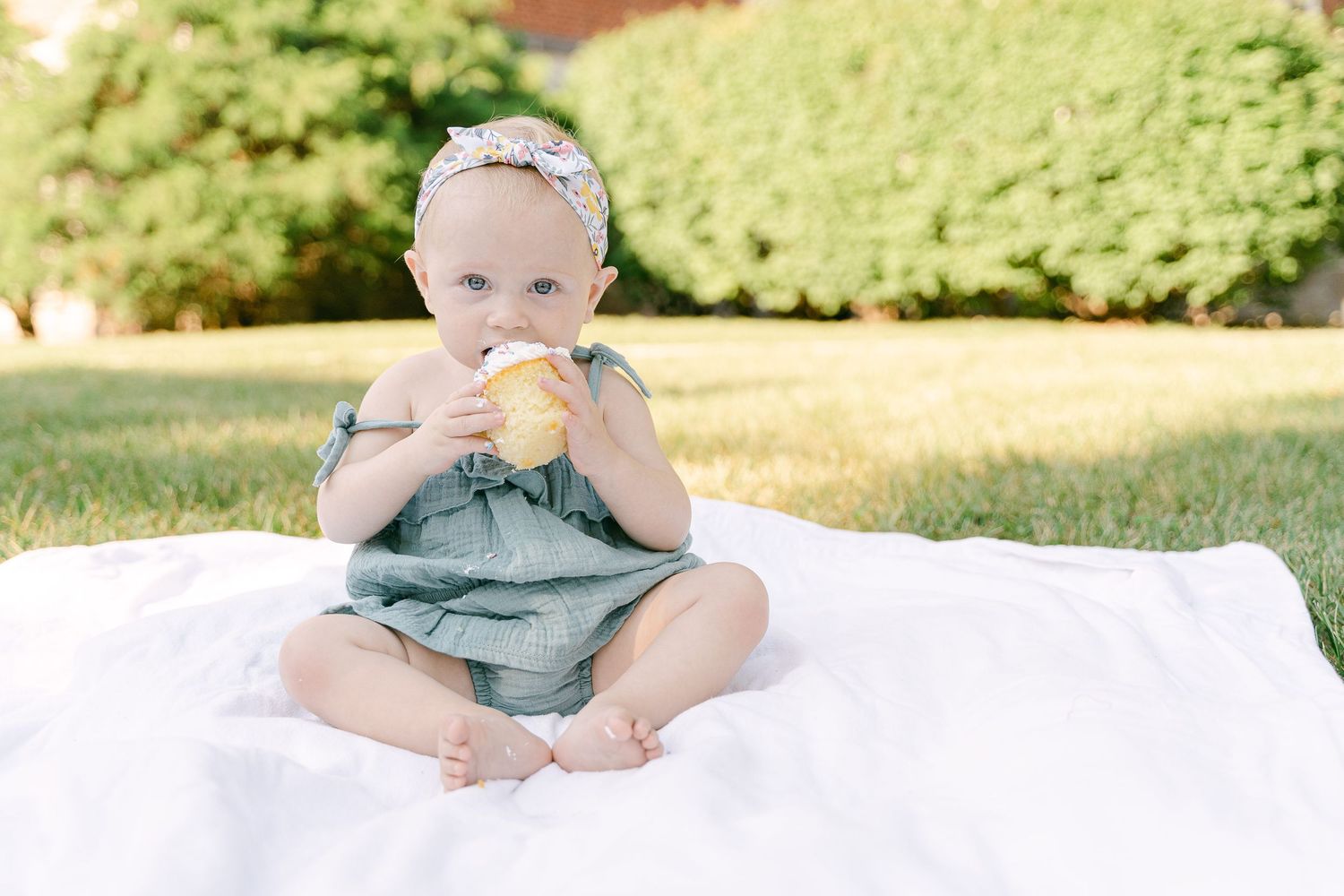 A baby in a gray shirt sits on a white blanket outdoors in the grass during a sunny day.