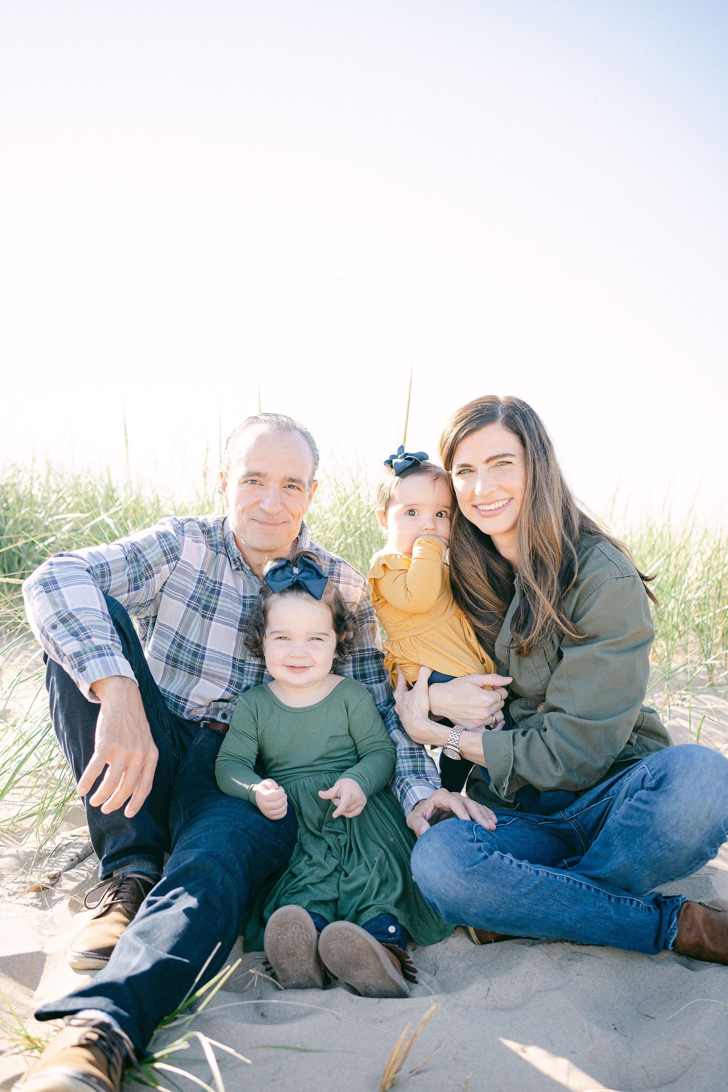 Family sits together on sandy beach during sunset photoshoot wearing casual clothing.