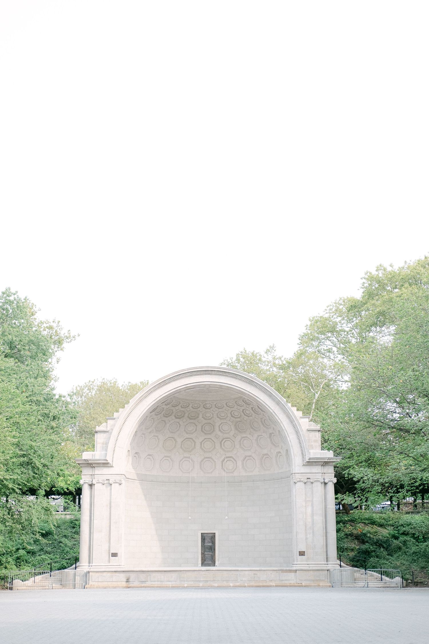 A panoramic view of a classic white stone bandshell or amphitheater in a park surrounded by green trees.