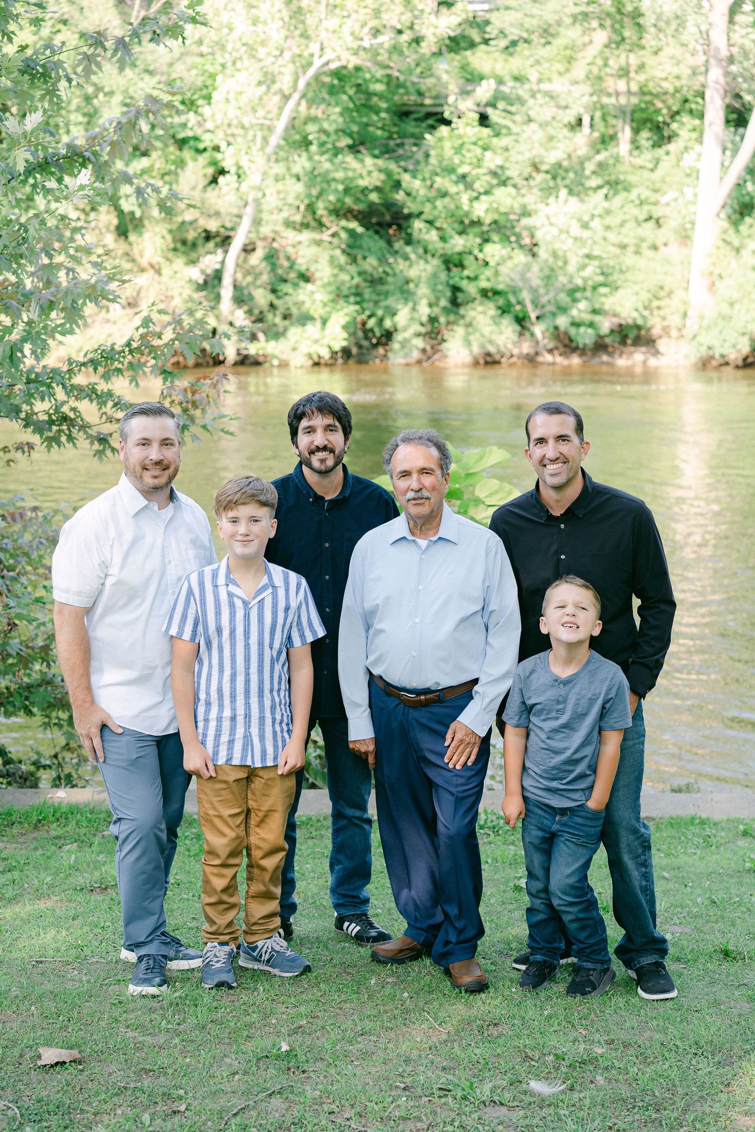 A multi-generational group stands together on grass near a river surrounded by lush trees.