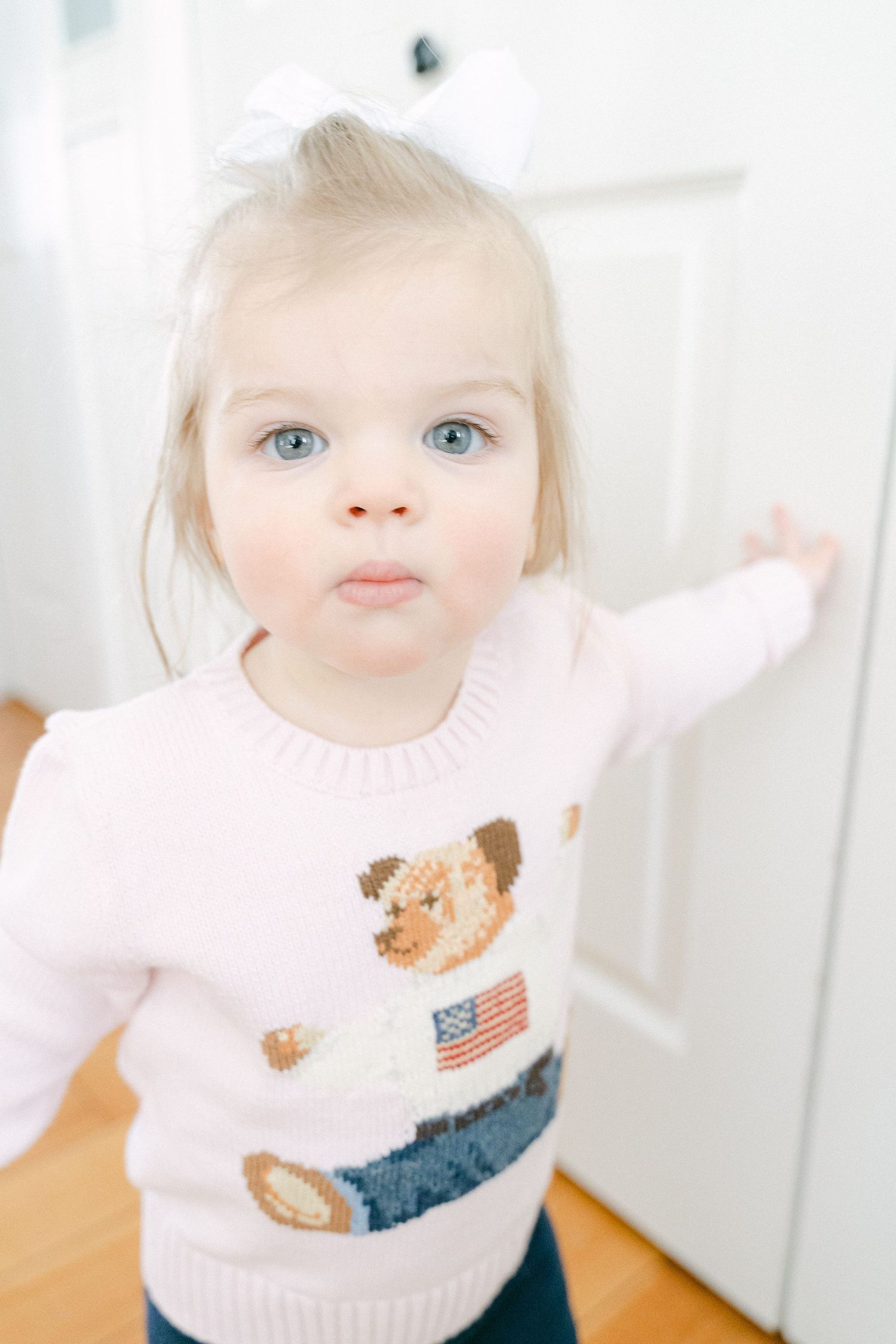 A toddler wearing a pink sweater with a teddy bear and American flag design stands in front of a white door.
