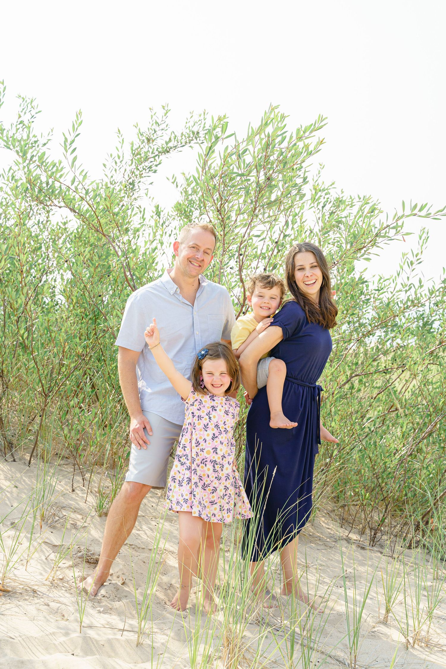 Family of four poses together among beach grass and sand dunes on a sunny day.