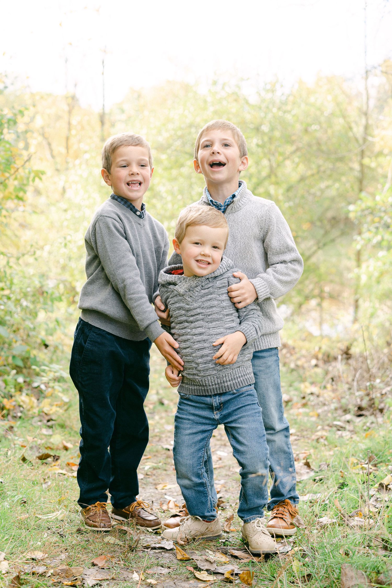 A playful outdoor photoshoot showing people in coordinated grey sweaters and dark pants on grass with autumn leaves.