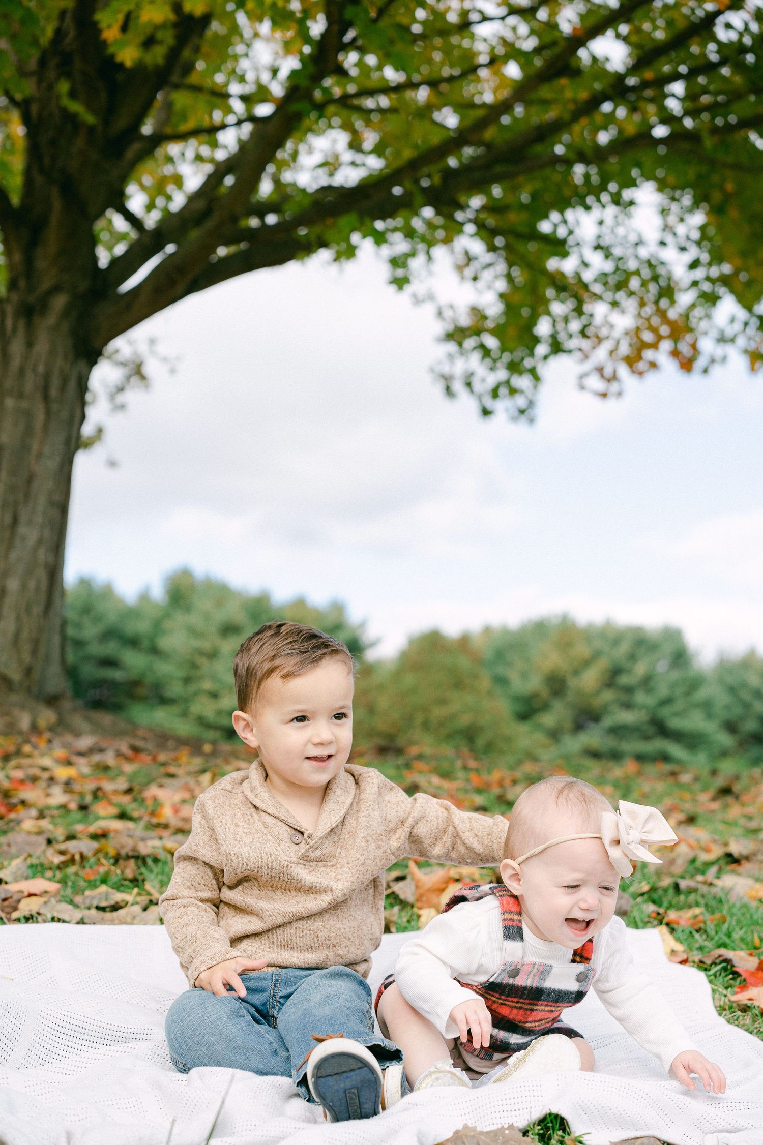 Two children in matching beige sweaters sit on a white blanket surrounded by autumn leaves in a park setting.