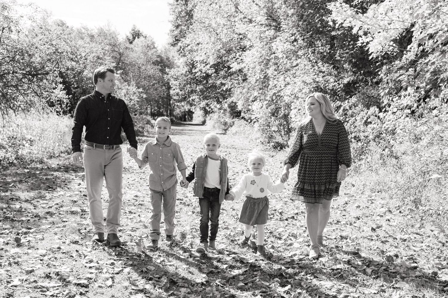 A group of people walk together along a wooded trail covered in fallen autumn leaves.