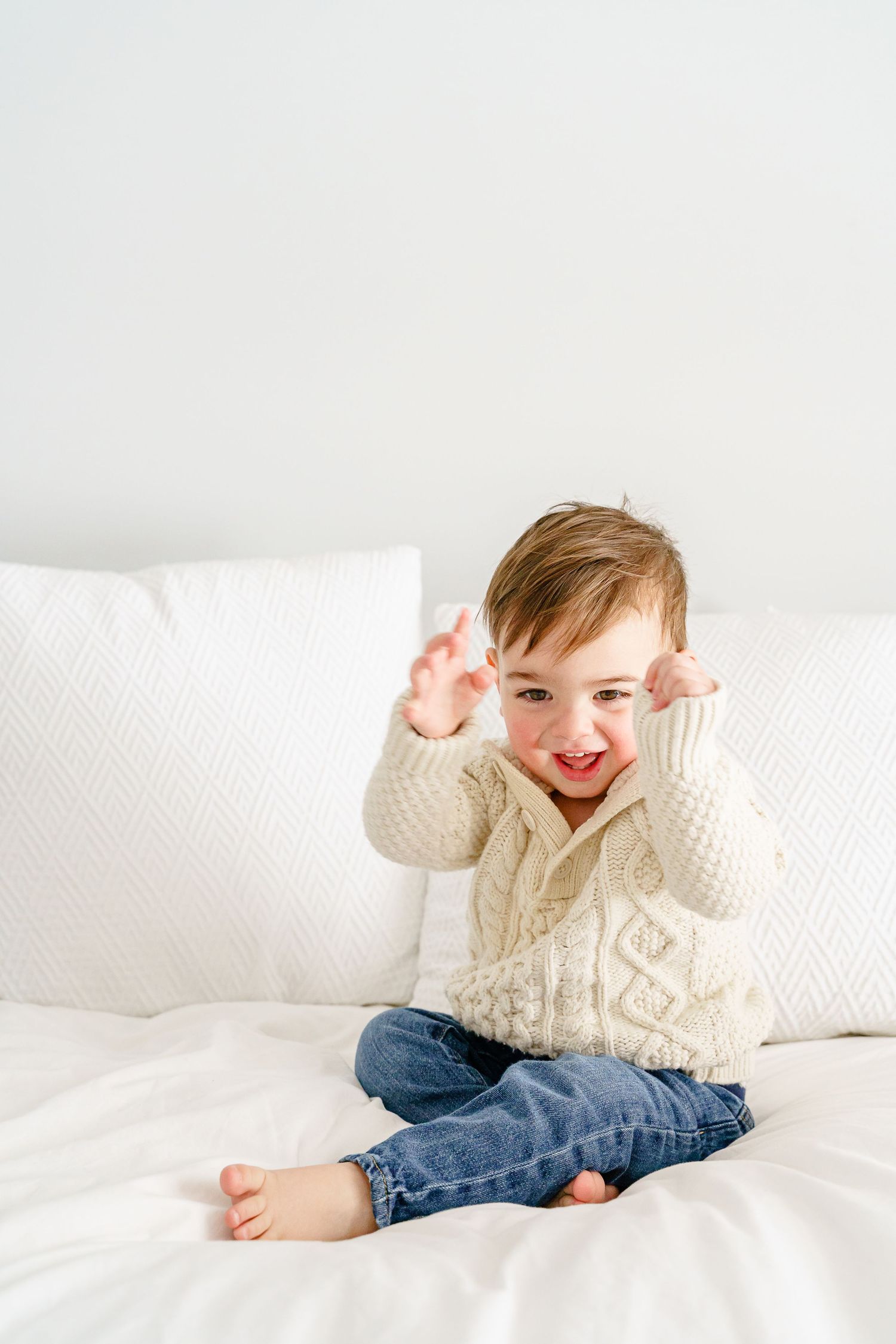 Child in cream knit sweater and jeans lying on white bedding while playing and smiling.