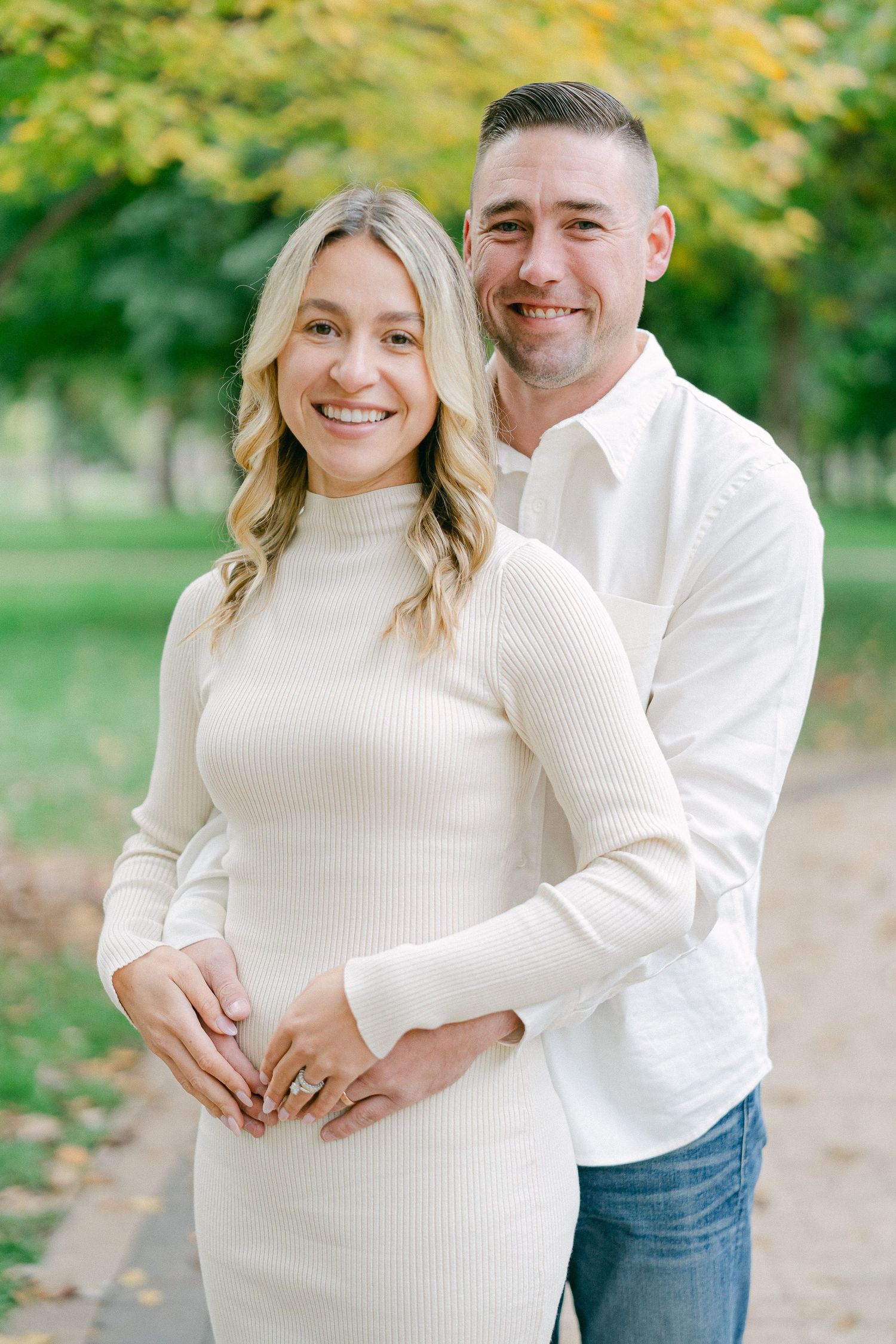 A couple in matching white sweaters share tender moments in a park with autumn foliage in the background.
