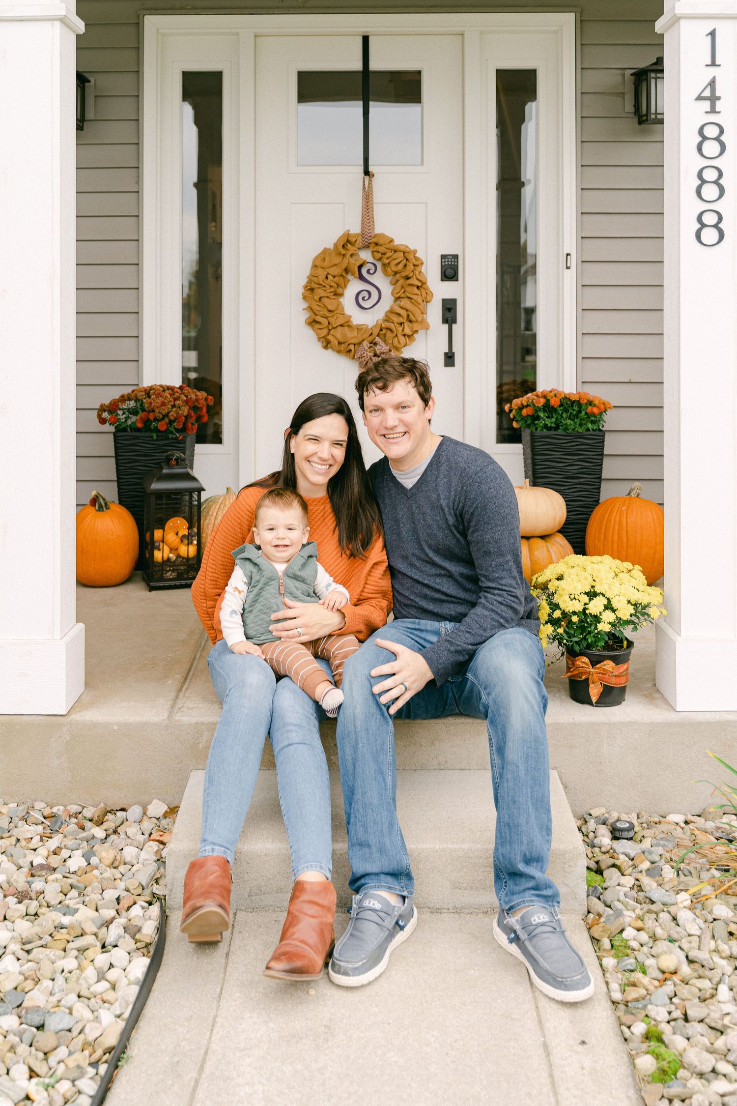 A family sits together on a front porch decorated with fall pumpkins and mums.