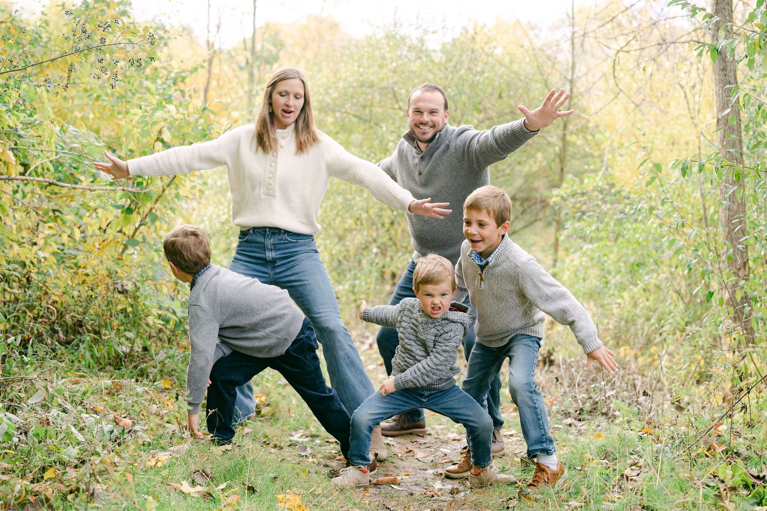 A playful family in casual attire frolics and poses together outdoors in a natural autumn setting.