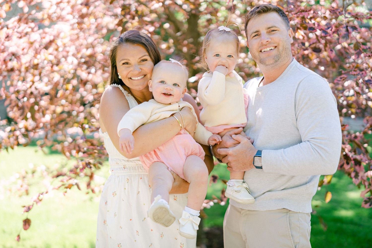 Family wearing coordinated white and neutral outfits poses together outdoors with pink flowering trees in background.