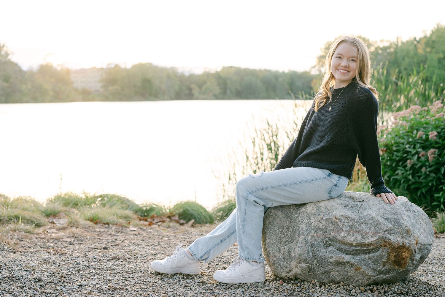 A person wearing a white dress sits gracefully on a large rock formation at sunset near the ocean.