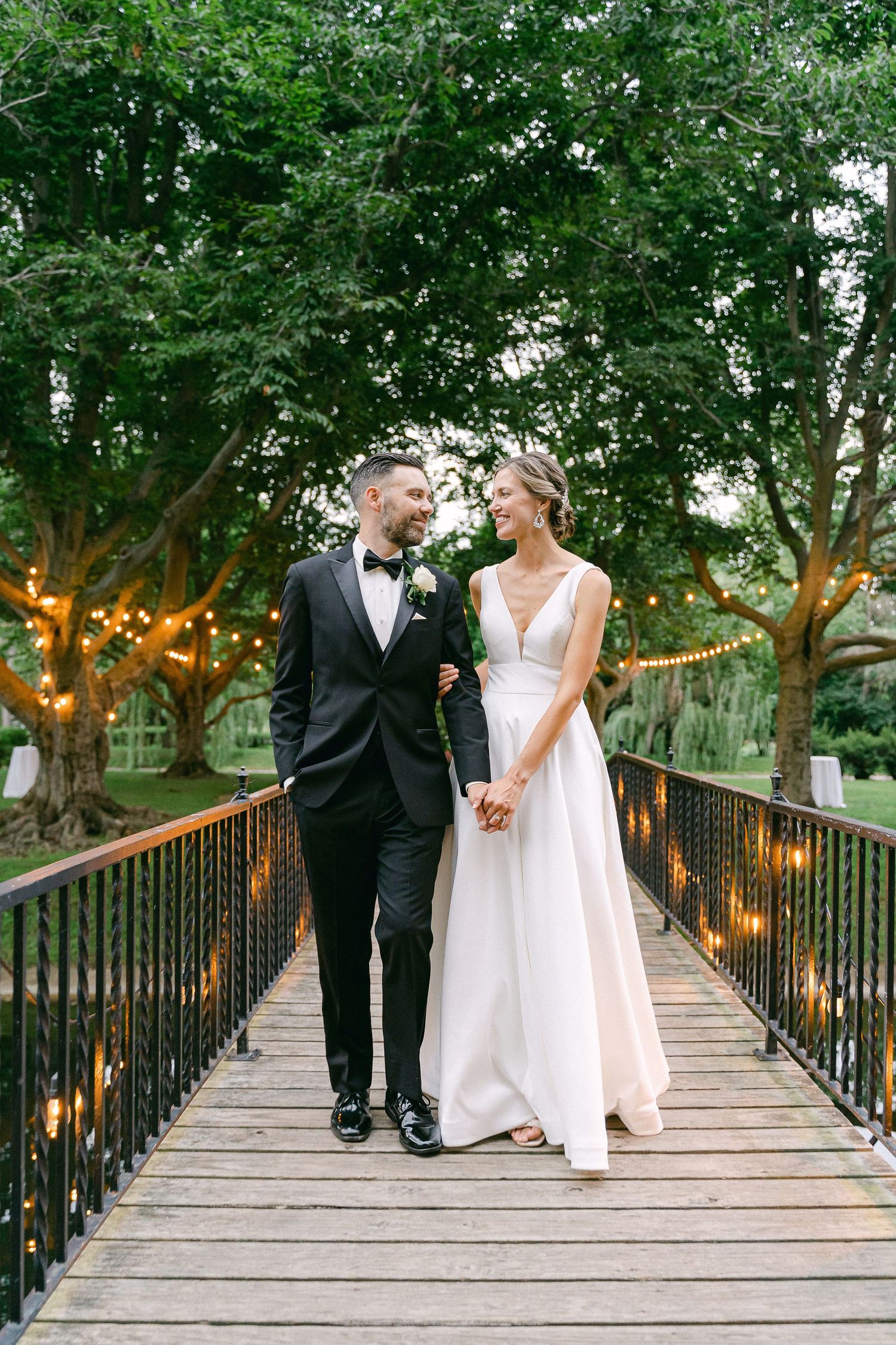 Bride and groom holding hands on a wooden bridge surrounded by lush trees and string lights at an outdoor wedding in South Bend, IN