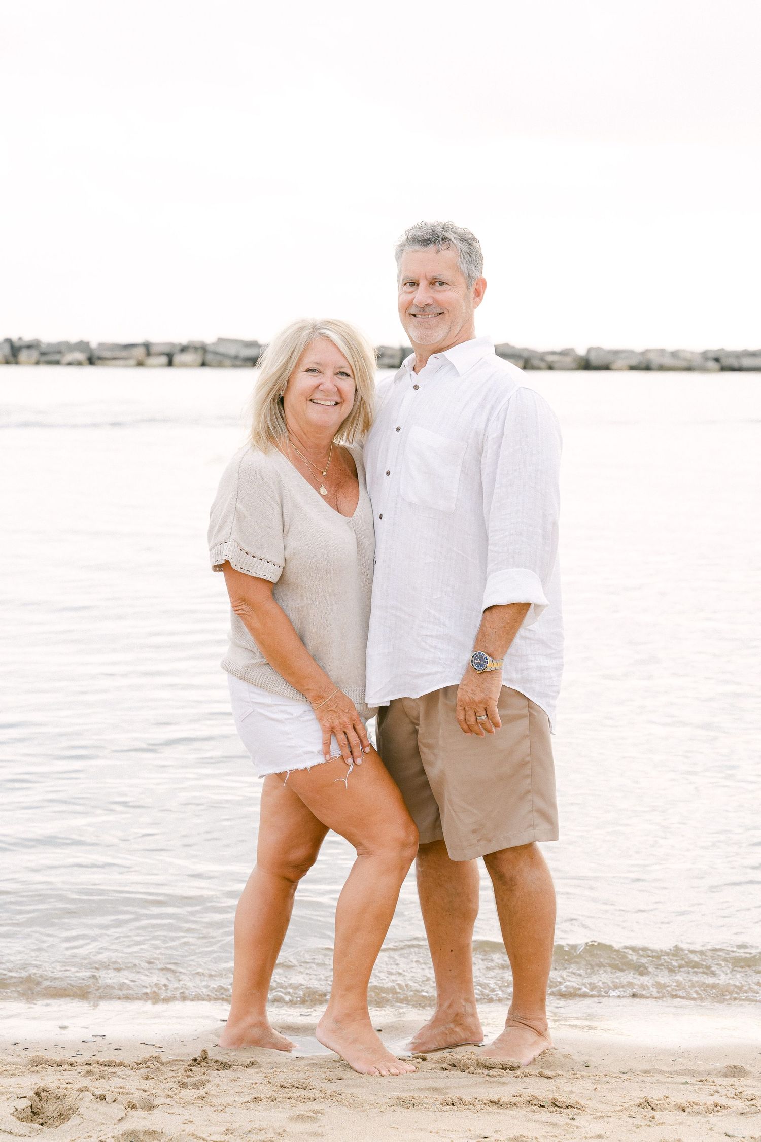 Couple poses together on sandy beach with water and bright sky in background.