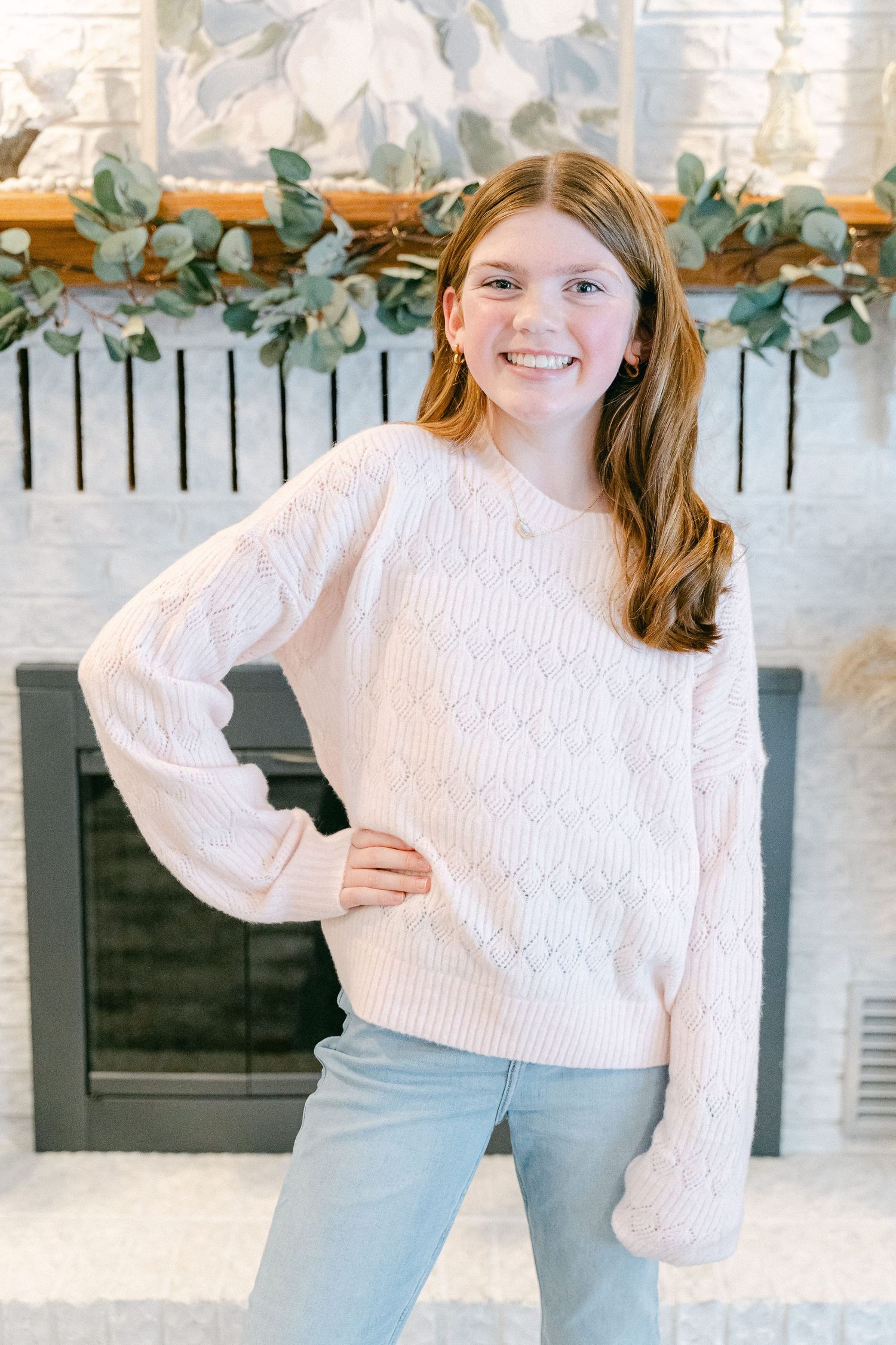 Person in a cozy white cable knit sweater poses against a white brick wall with eucalyptus garland decor.