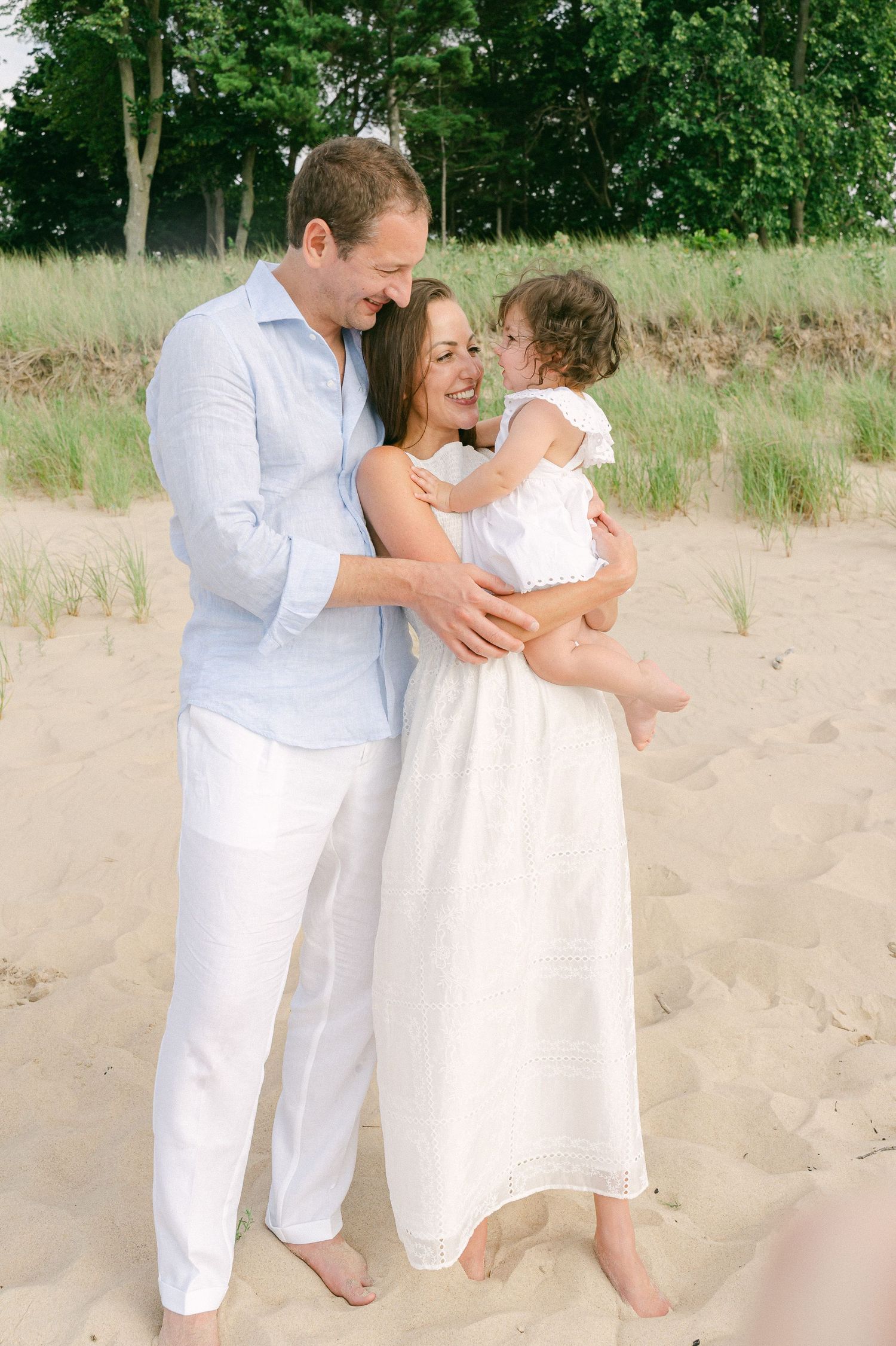 A family shares tender moments together on a sandy beach while wearing all white outfits.