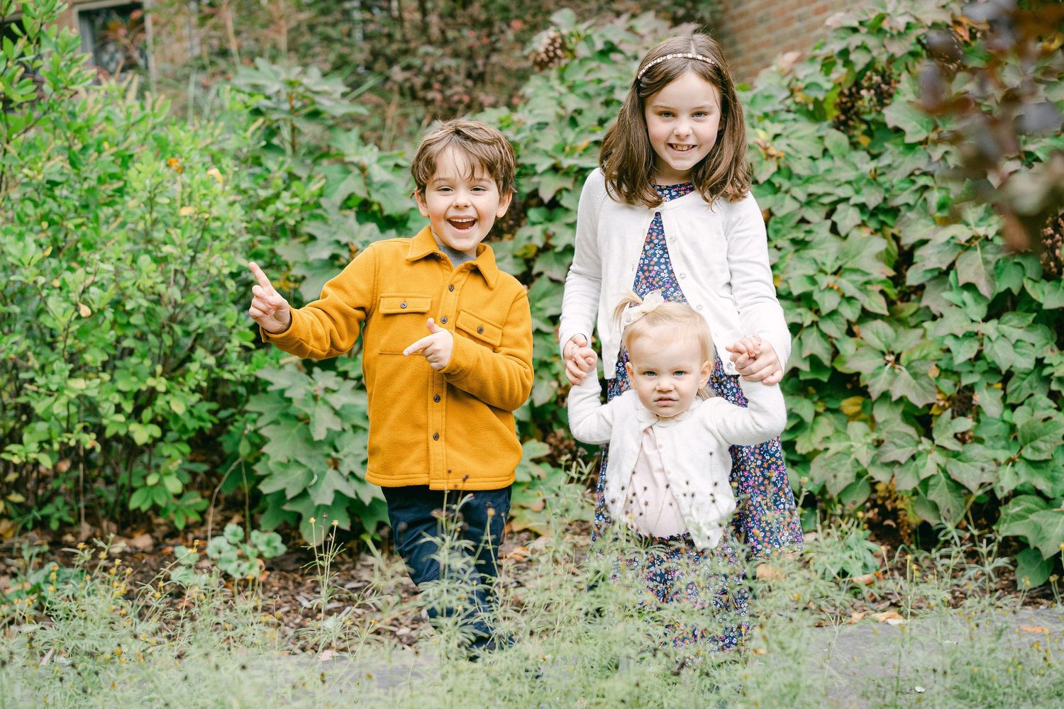 Three children stand together outdoors among green foliage, wearing casual fall clothing.