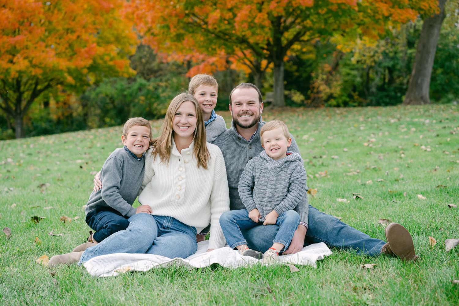 A family group sits together on grass during autumn with colorful fall foliage in the background.