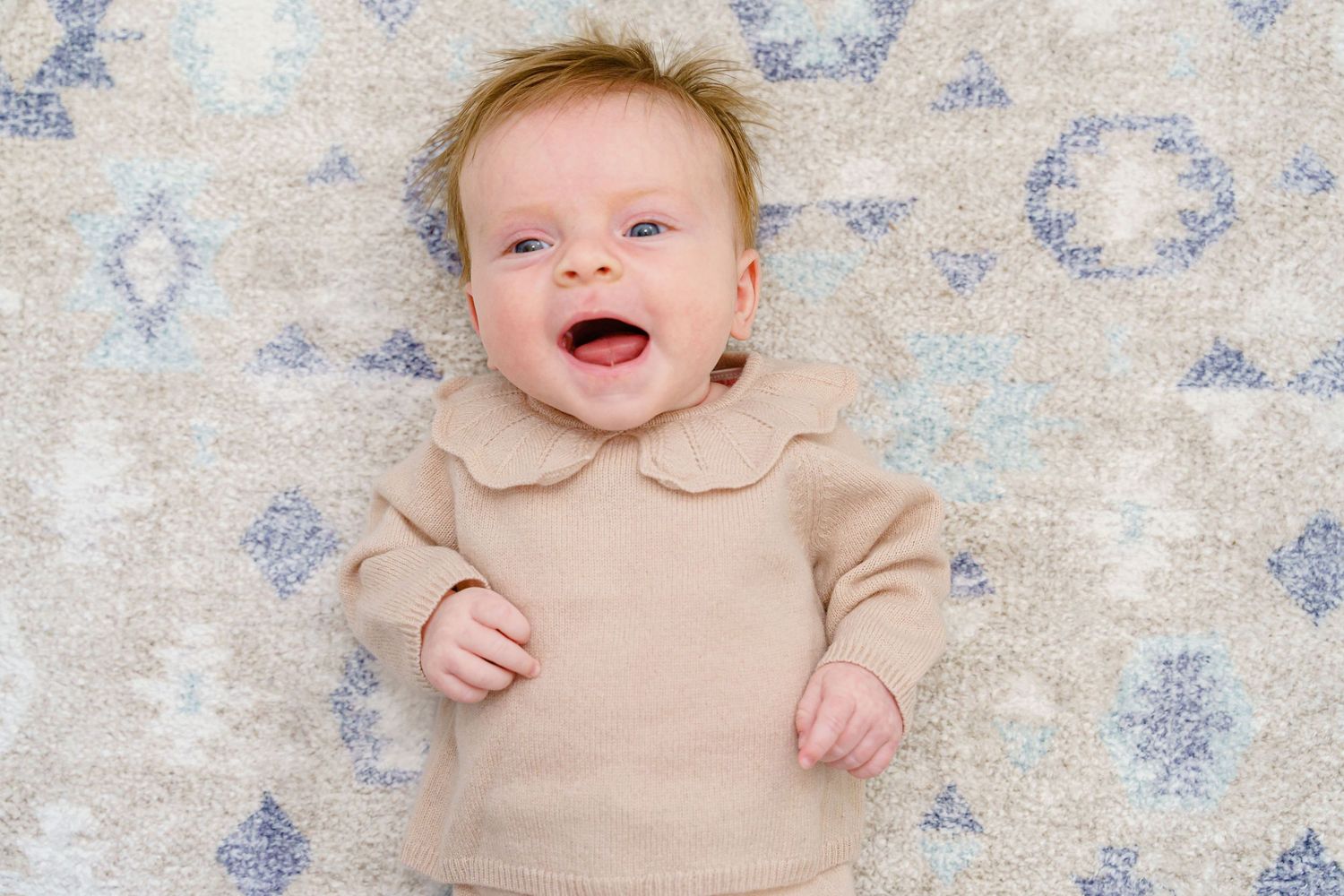 A smiling baby wearing a beige onesie lies on a patterned blue and white rug.