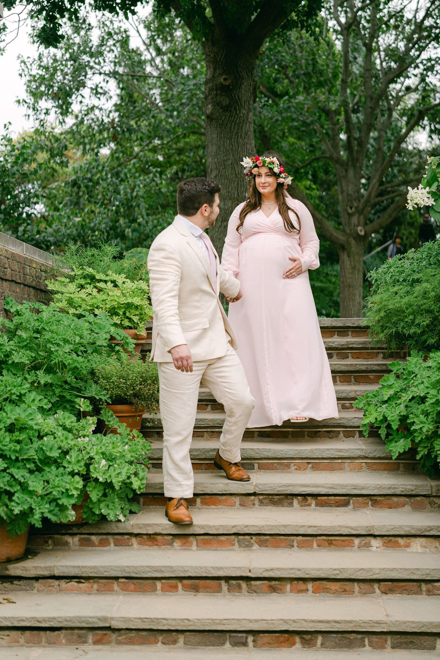A couple in pastel outfits stand together on brick garden steps surrounded by lush greenery.