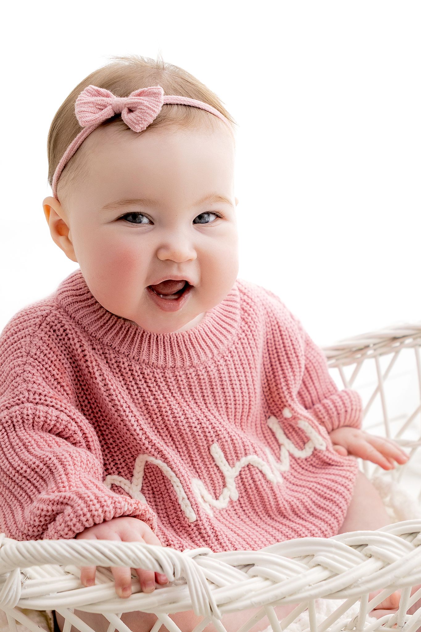 Cheerful baby girl in pink knit sweater sitting in white wicker basket smiling.