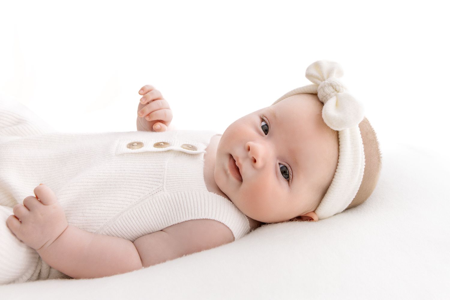 Adorable baby in white outfit and bow headband lying on white background.
