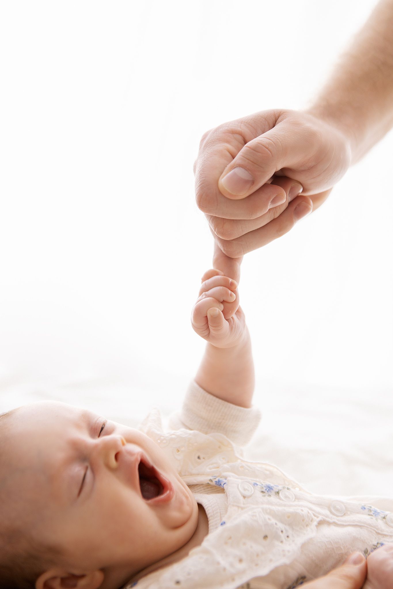 A crying newborn baby holds an adult finger while lying on a white surface in a tender bonding moment.