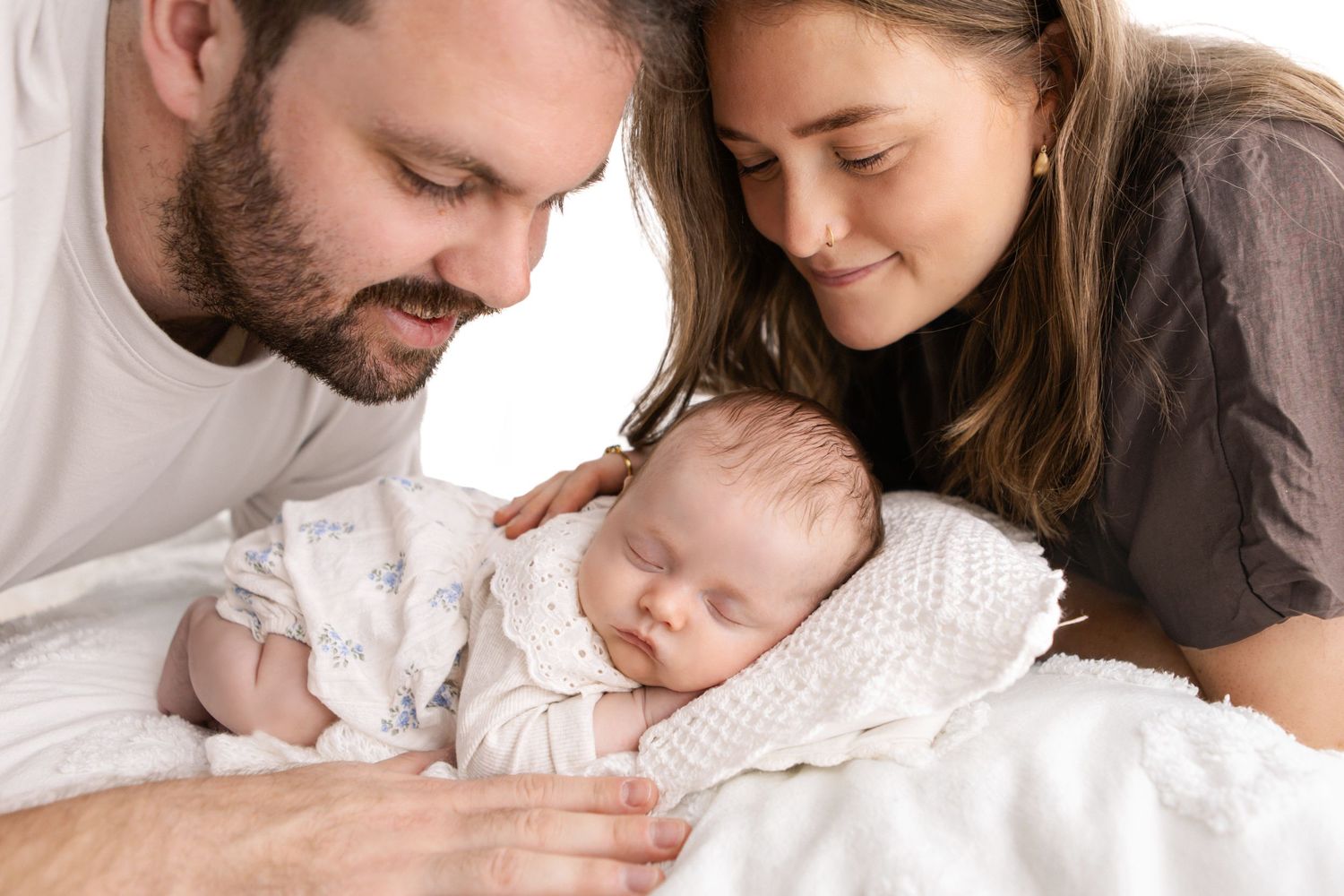 A loving couple gazes adoringly at their sleeping newborn baby wrapped in a white knit blanket.