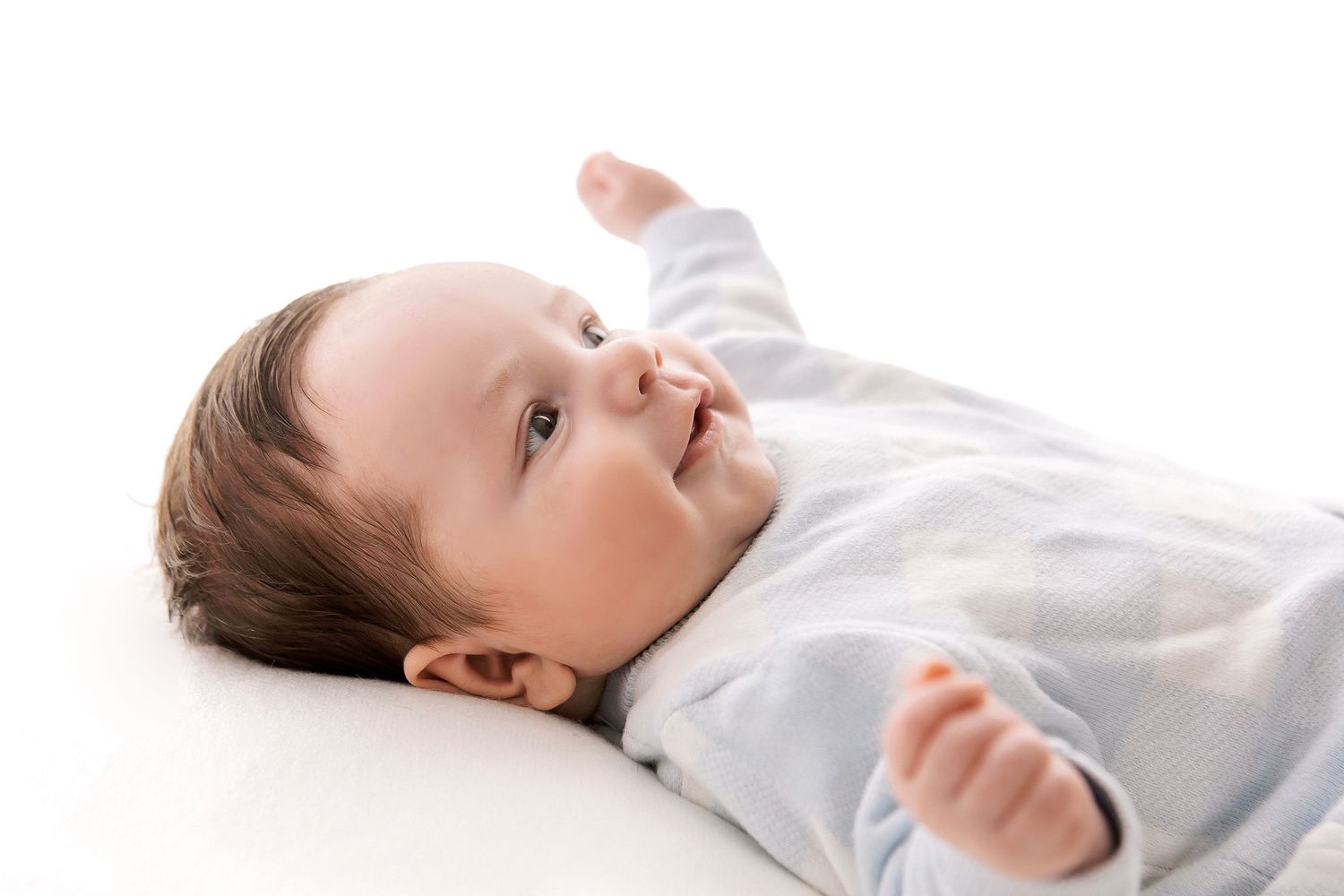 Cute baby boy lying on back in blue onesie, smiling and reaching up with arm on white background.