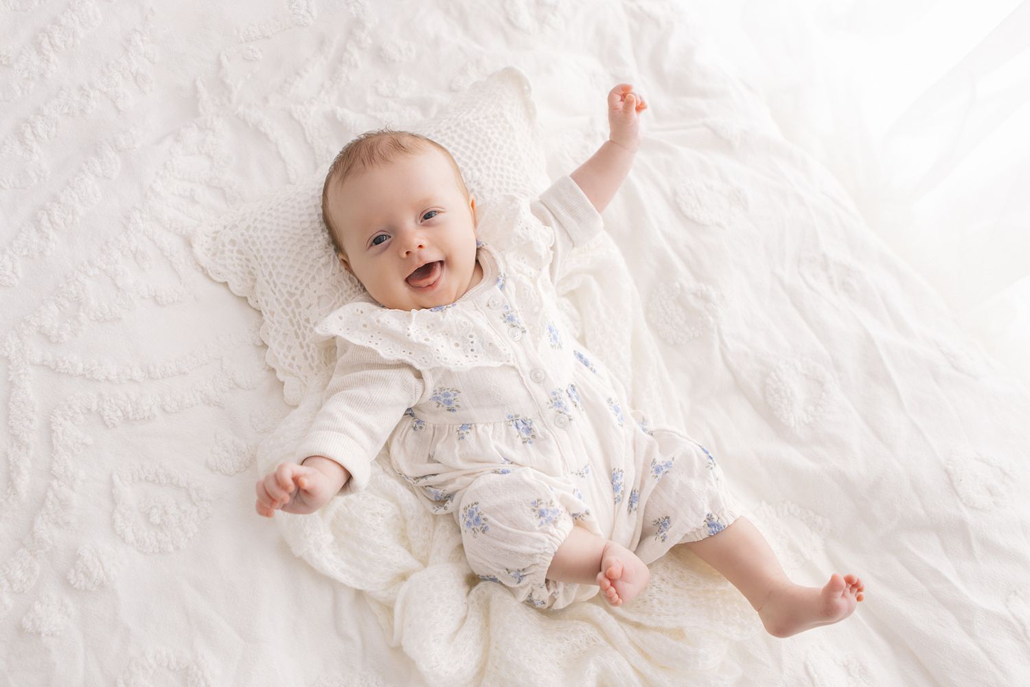 Smiling baby lying on white bedding wearing a floral romper with one arm raised, captured in a bright airy nursery photo.