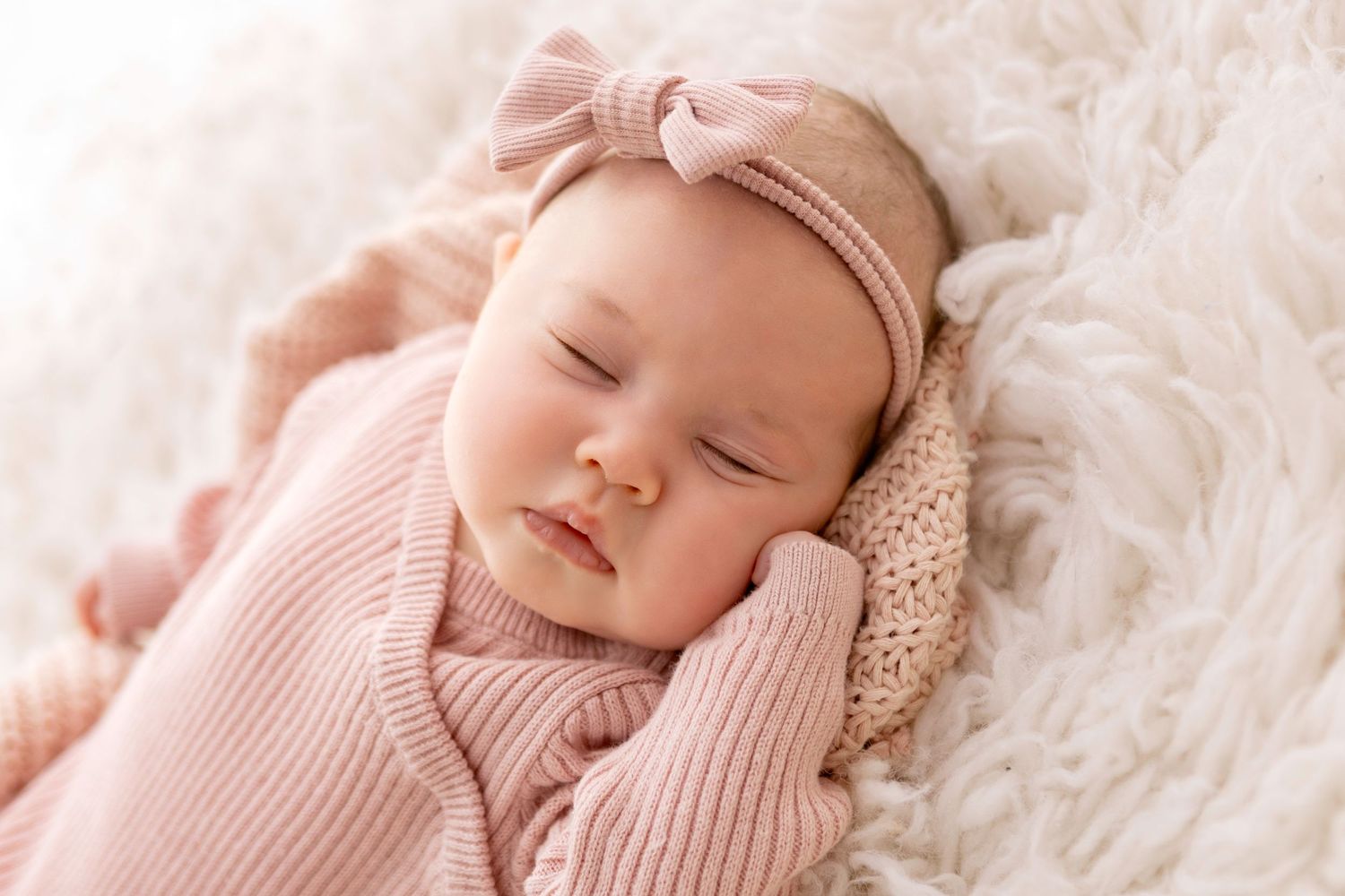 Sleeping newborn baby girl in pink knit swaddle and bow headband resting on fluffy white blanket.