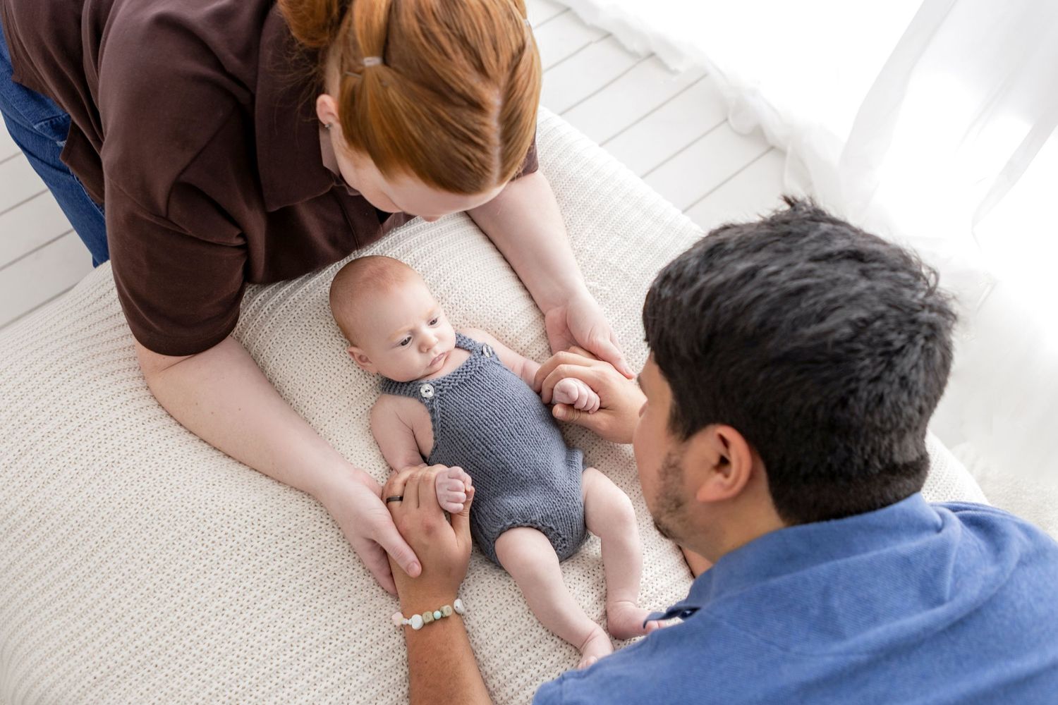 Two parents lovingly interact with their newborn baby lying on a soft white blanket at home.
