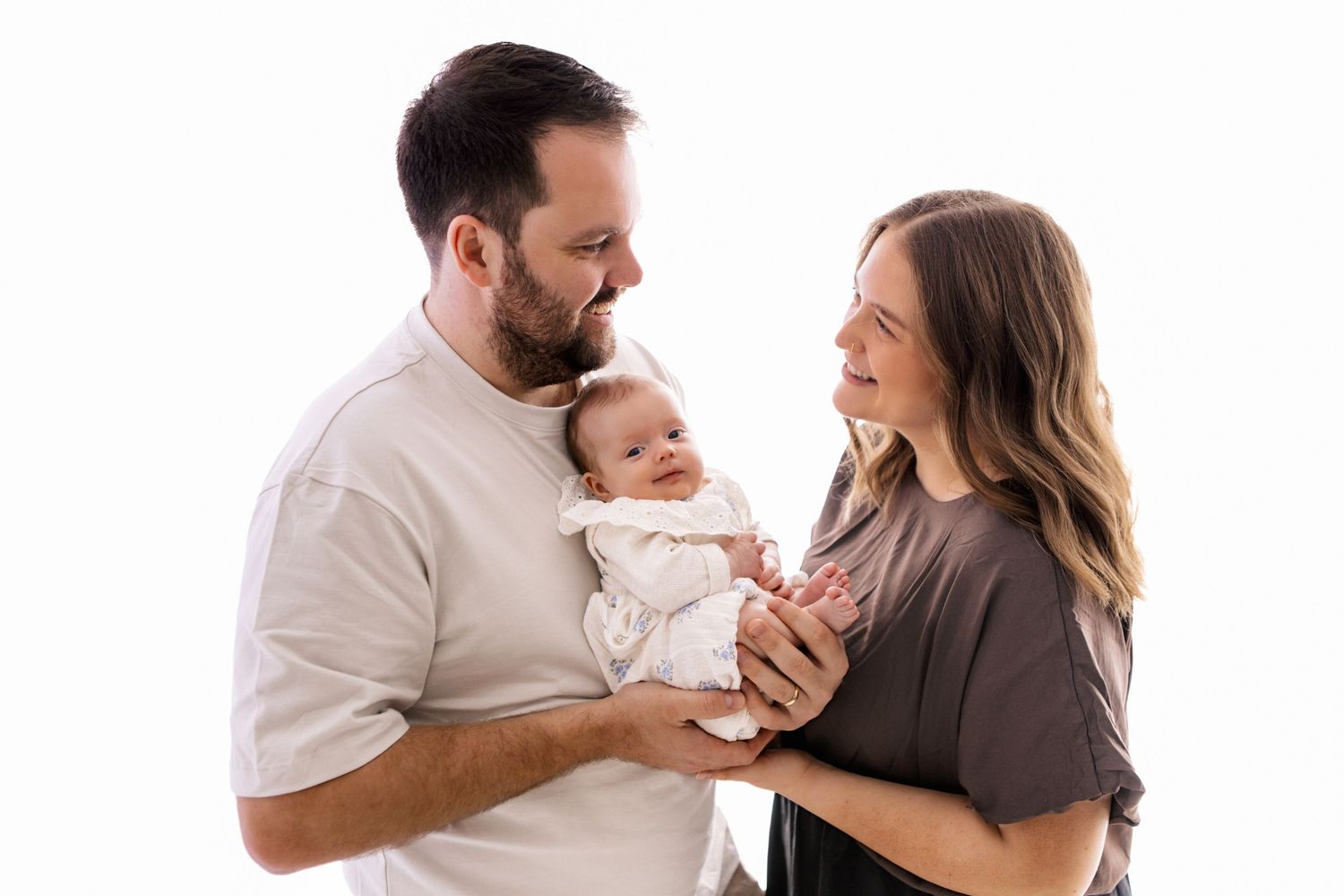 A couple smiles lovingly at their newborn baby during a professional white background studio portrait session.