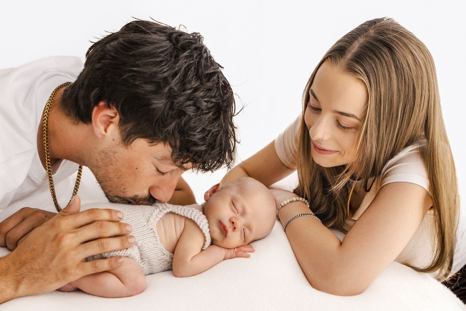 Parents lovingly kissing and gazing at their sleeping newborn baby on a white background.