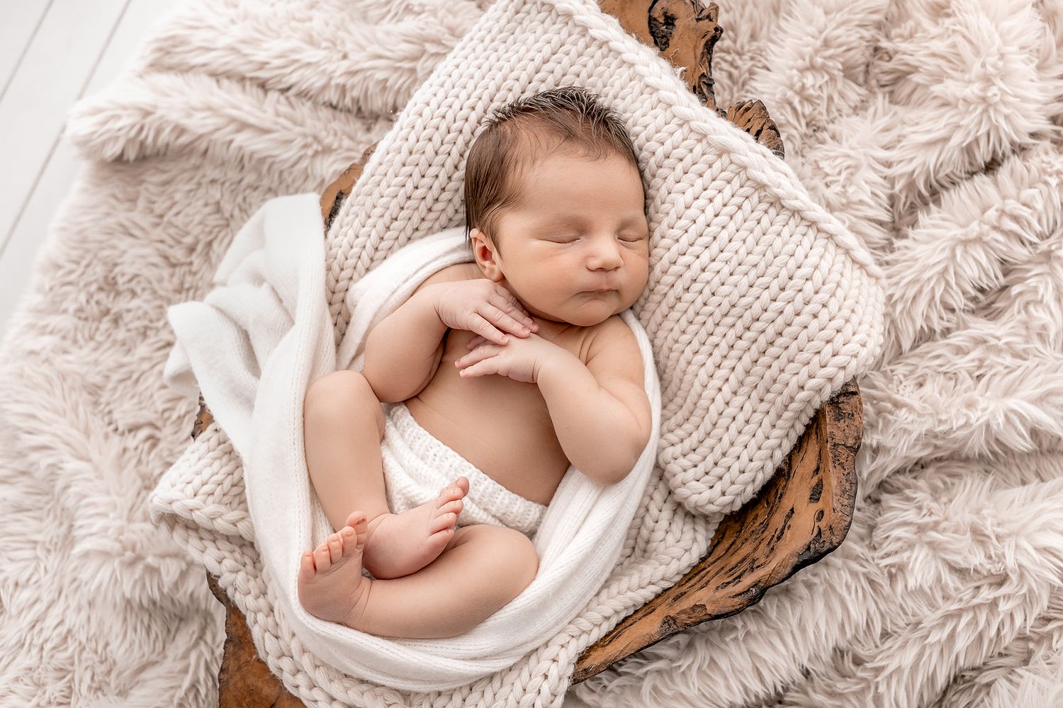 Newborn baby sleeping peacefully in a basket with soft beige knit blankets in a cozy newborn photography setup.
