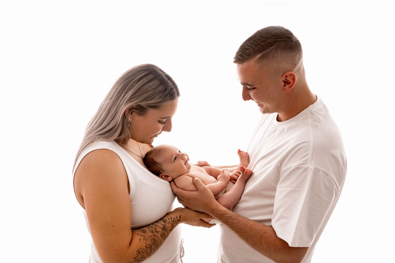 A couple in white outfits tenderly holding their newborn baby together against a white background.
