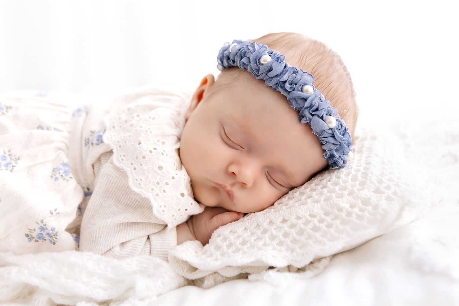 Peaceful newborn baby sleeping on white blanket wearing a delicate blue floral headband in soft natural light.