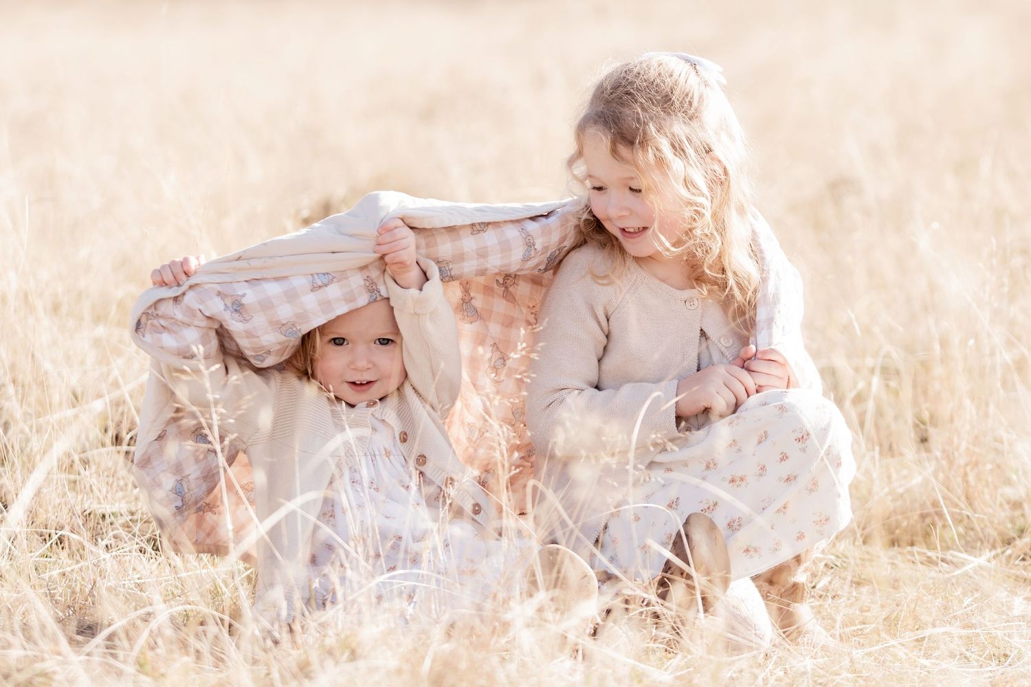 Two young children playing under a blanket in a golden dry grass field outdoors.