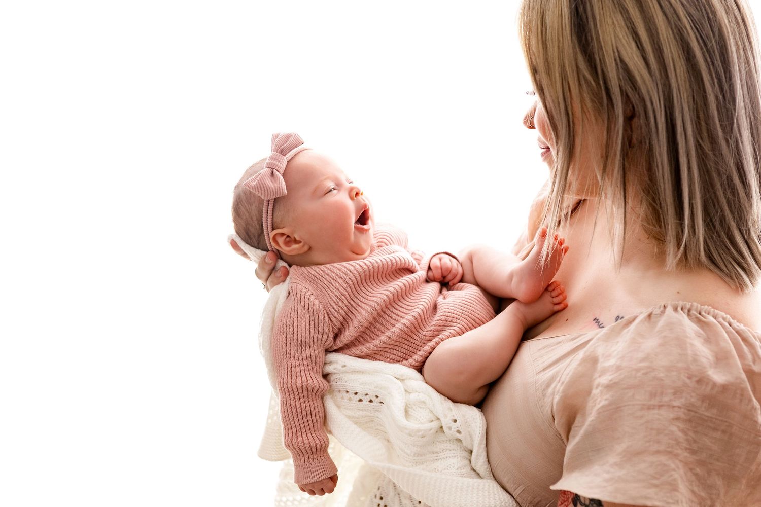Mother holding laughing newborn baby girl in pink knit outfit against white background.