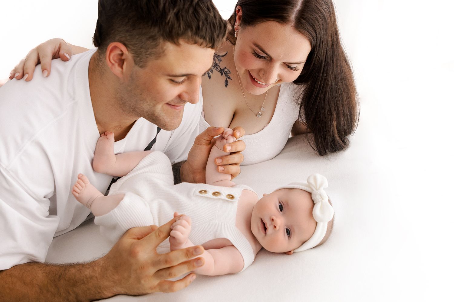A happy couple in white outfits lying down and interacting with their smiling baby girl wearing a white bow headband.