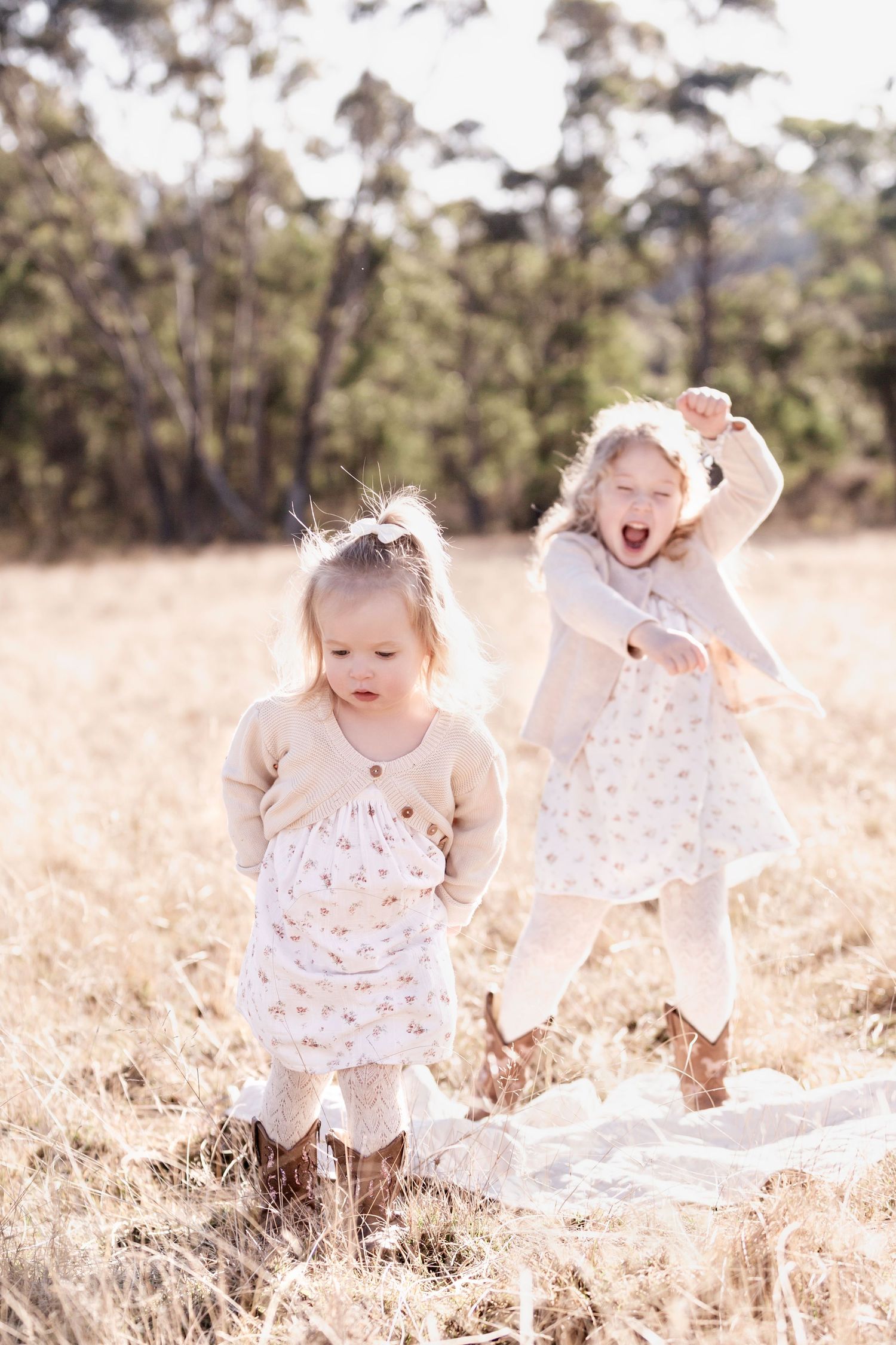 Two young girls in floral dresses playing energetically in a sunlit golden field.