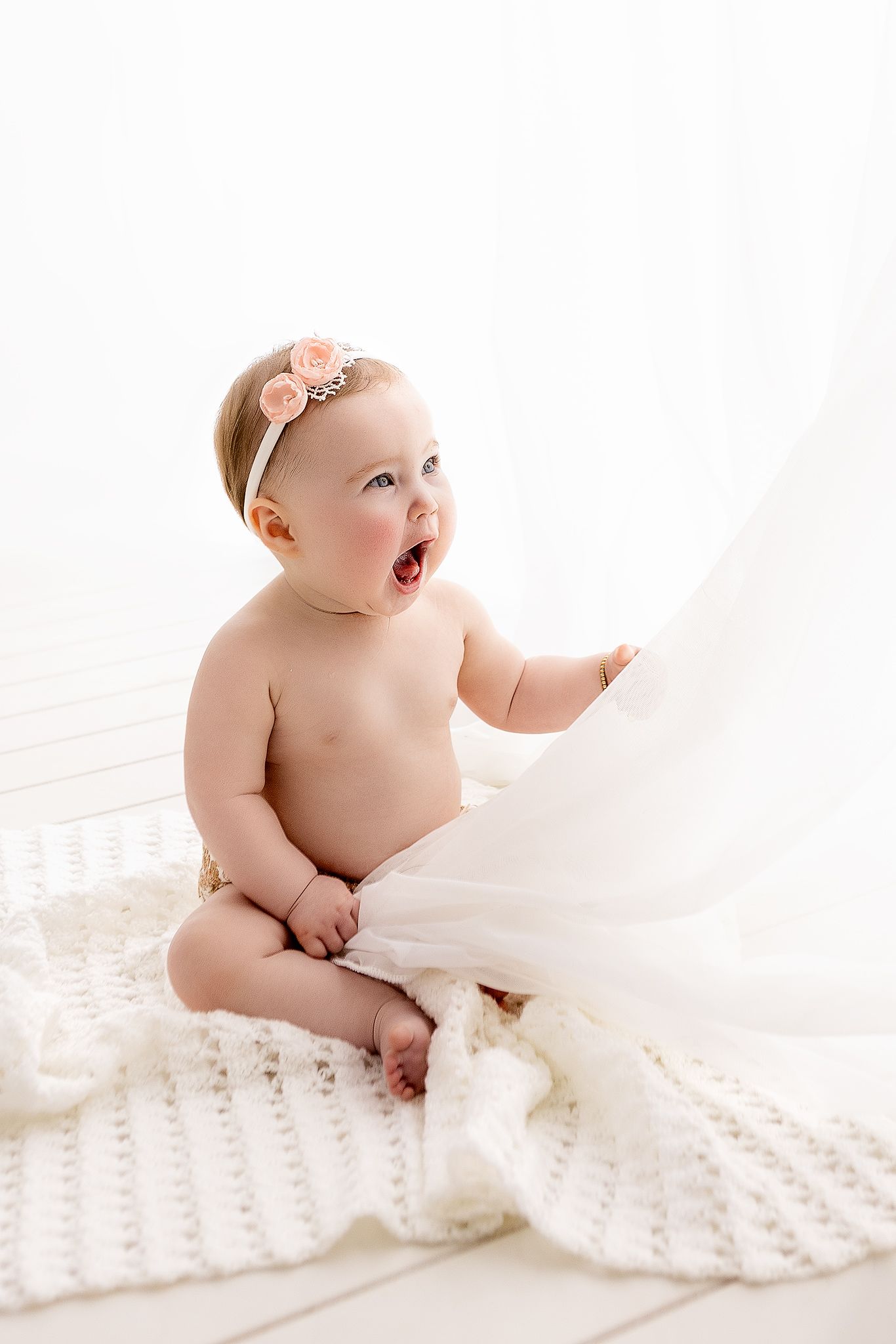 Curious baby girl in diaper with floral headband sitting on white knit blanket.