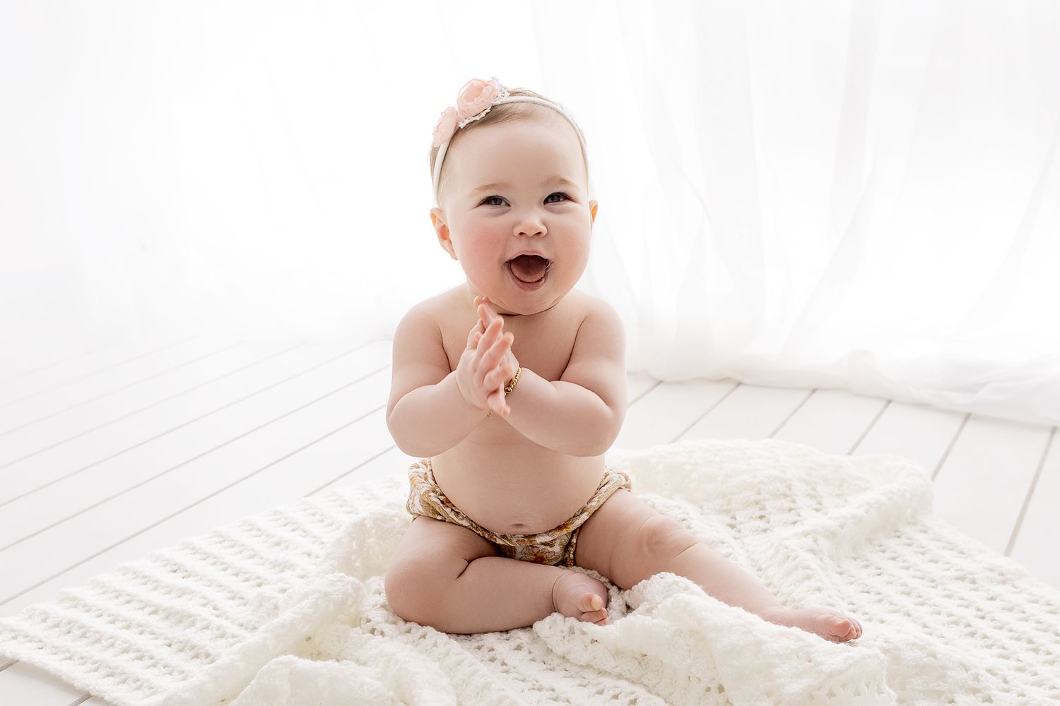 Happy baby girl in gold bloomers with pink bow headband clapping hands on white blanket.