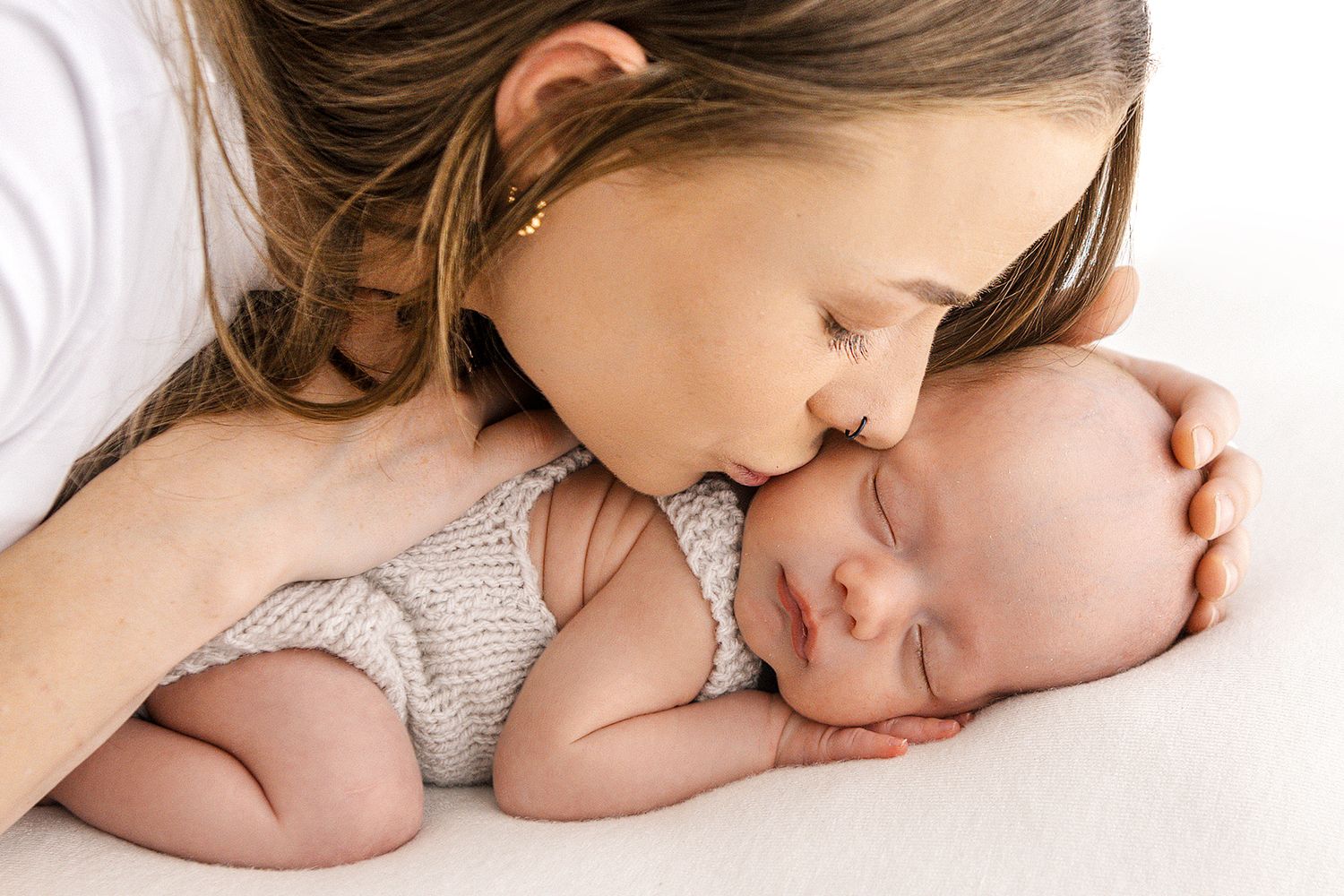 A young girl tenderly kisses a sleeping newborn baby on the head during a professional newborn photography session.