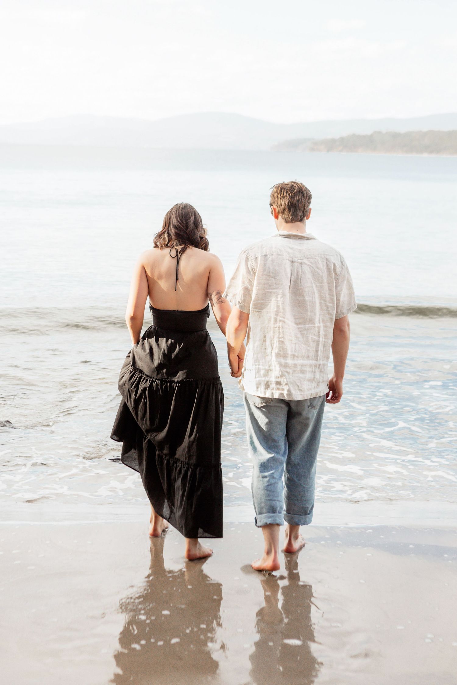 Couple holding hands walking barefoot on a sandy beach toward the ocean, woman in black dress, man in white shirt and jeans.
