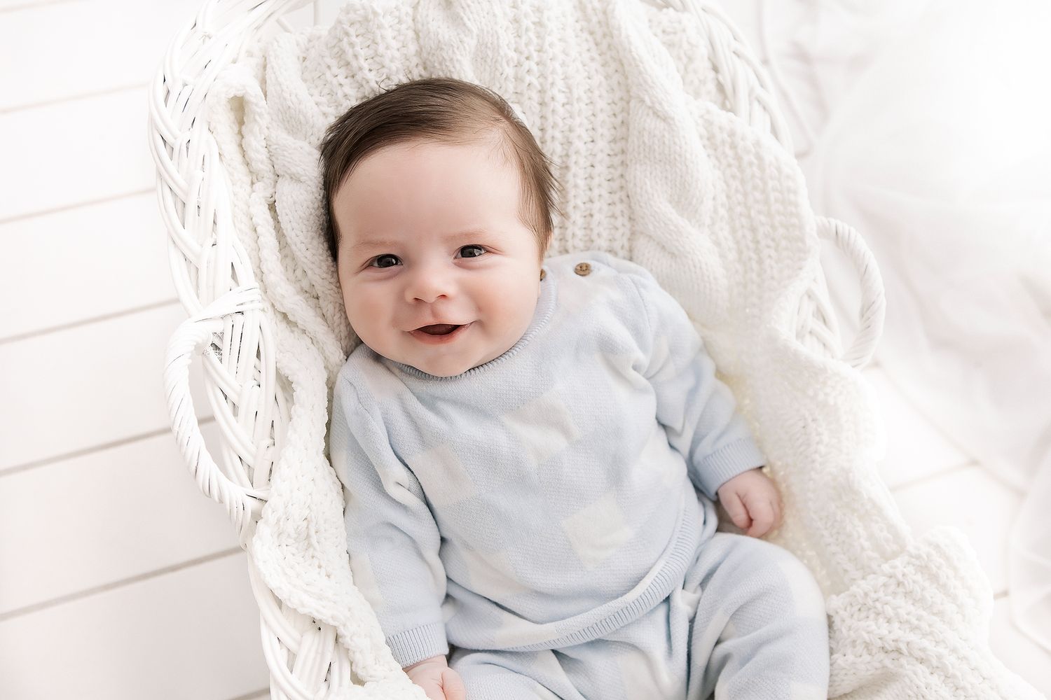 Smiling baby in light blue onesie resting in a white wicker chair with a cozy knit blanket.